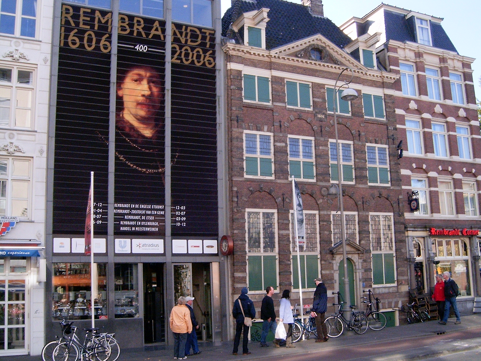La fachada del Museo Casa de Rembrandt con una gran pancarta de Rembrandt que abarca de 1606 a 2006, ventanas verdes y gente caminando afuera con bicicletas.