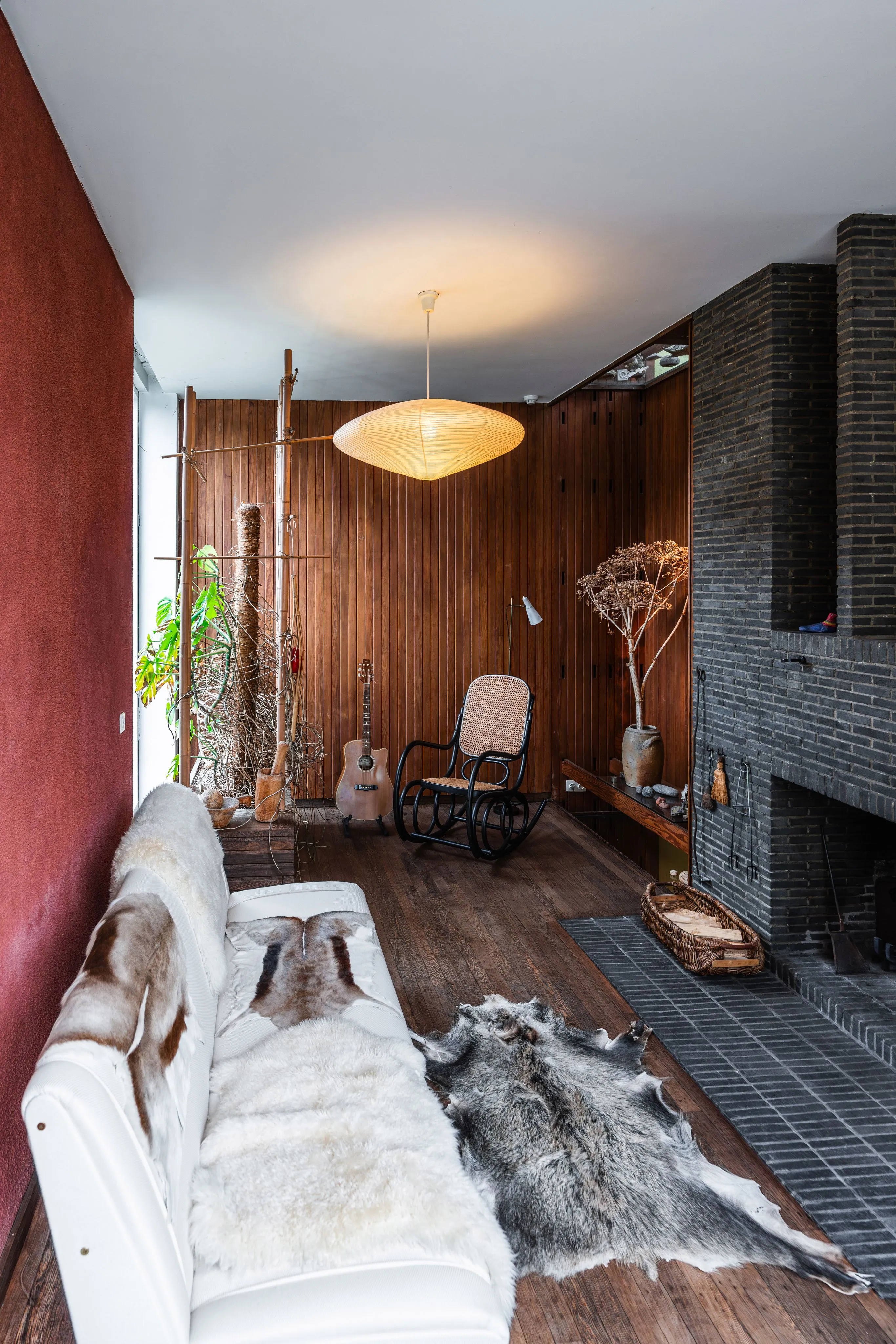 Interior de la Casa Renaat Braem, que presenta paredes revestidas de madera, una chimenea de ladrillo oscuro, un sofá blanco cubierto con pieles de animales y una lámpara colgante iluminada.