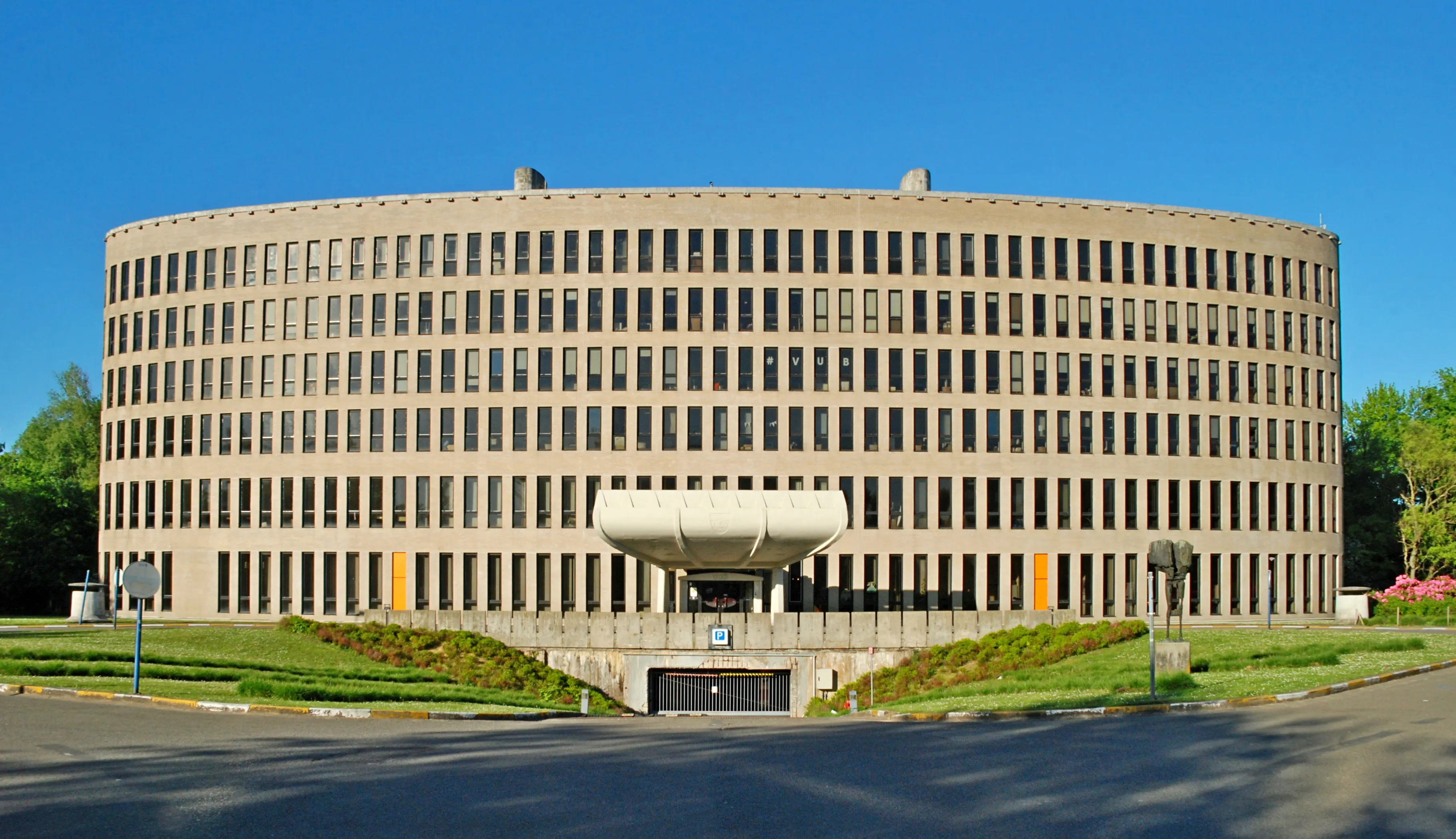 Toma exterior amplia de un gran edificio modernista de hormigón curvado con filas uniformes de ventanas y una marquesina de entrada central bajo un cielo azul despejado.
