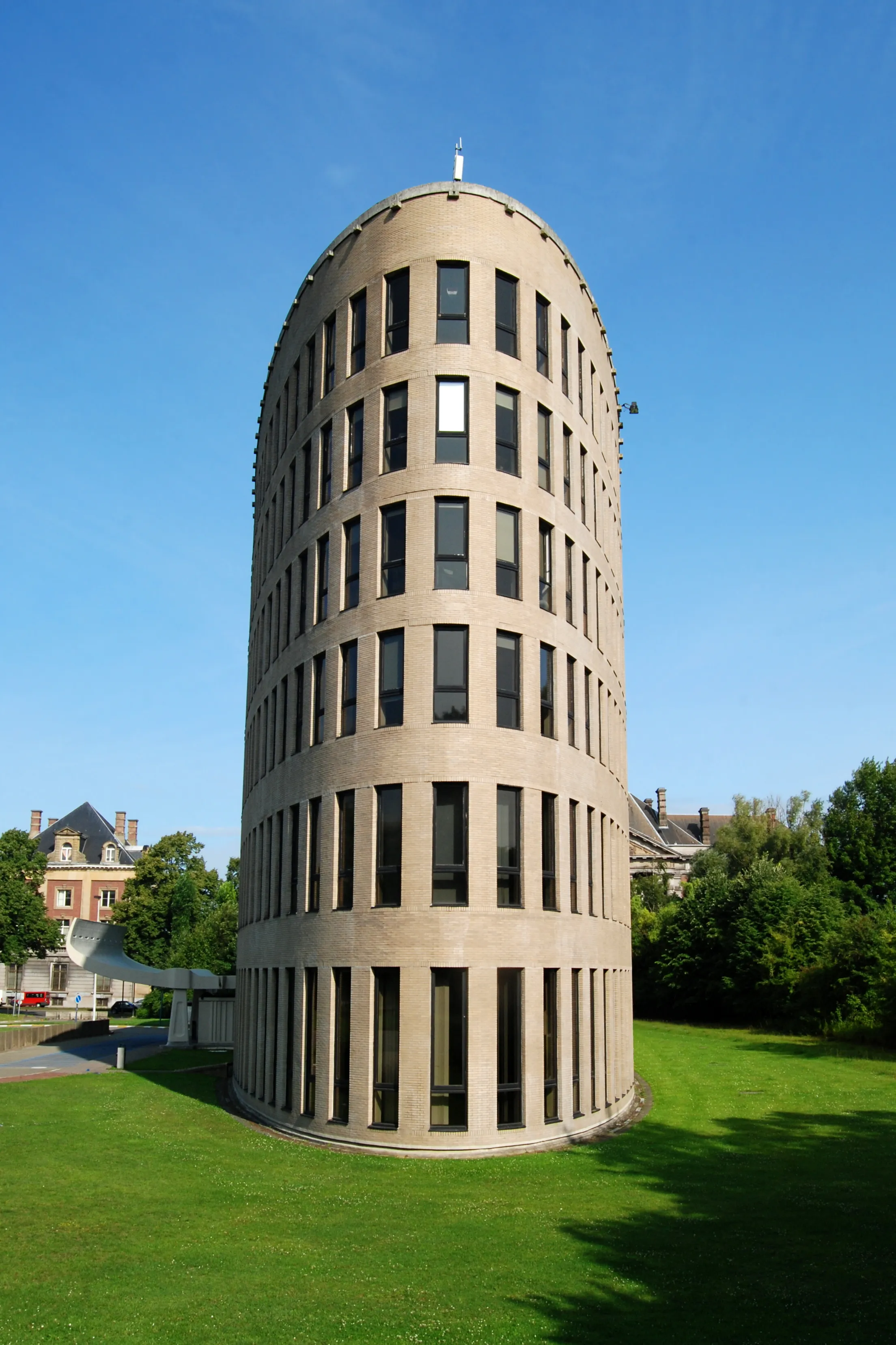 Alto edificio de torre modernista cilíndrica de ladrillo con franjas de ventanas verticales sobre un césped verde bajo un cielo azul despejado