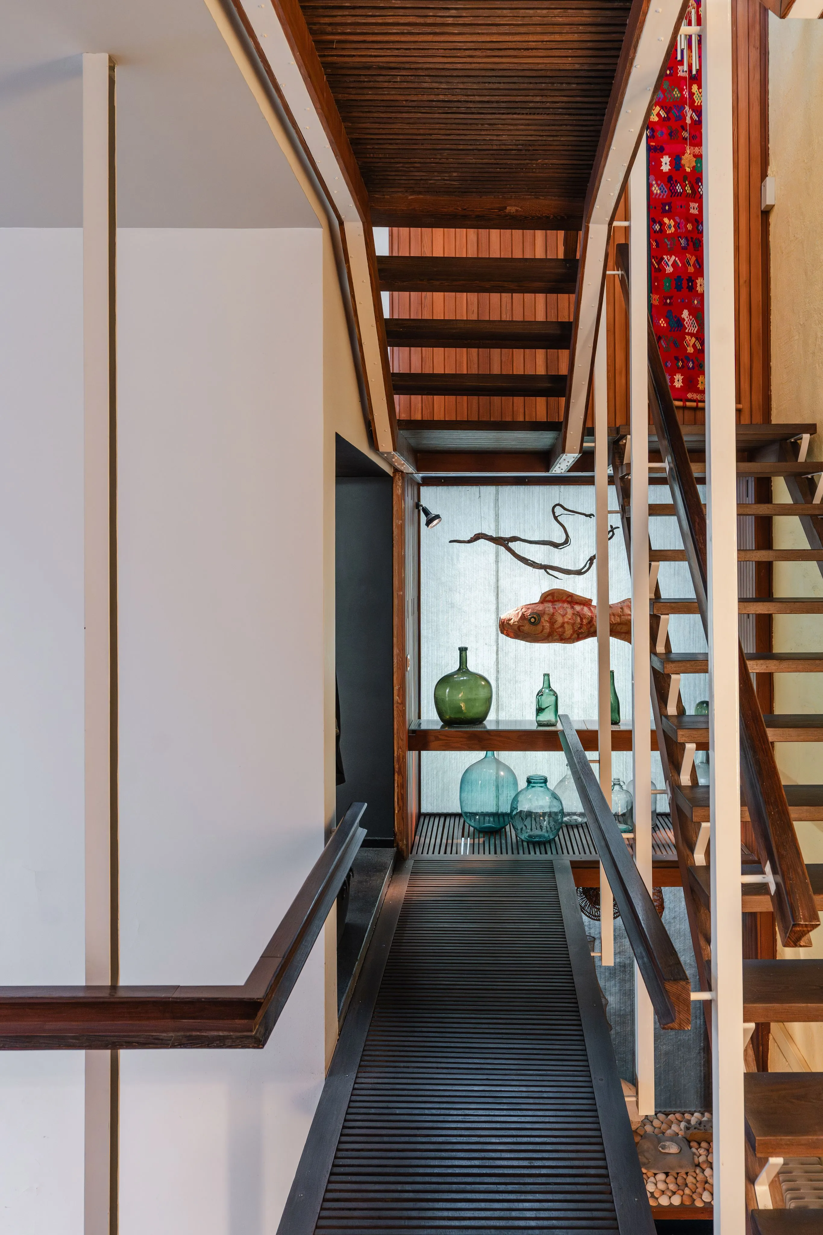 Vista interior de una casa modernista que presenta una escalera de madera, un techo de listones y una exposición de botellas de vidrio y una escultura de pared.