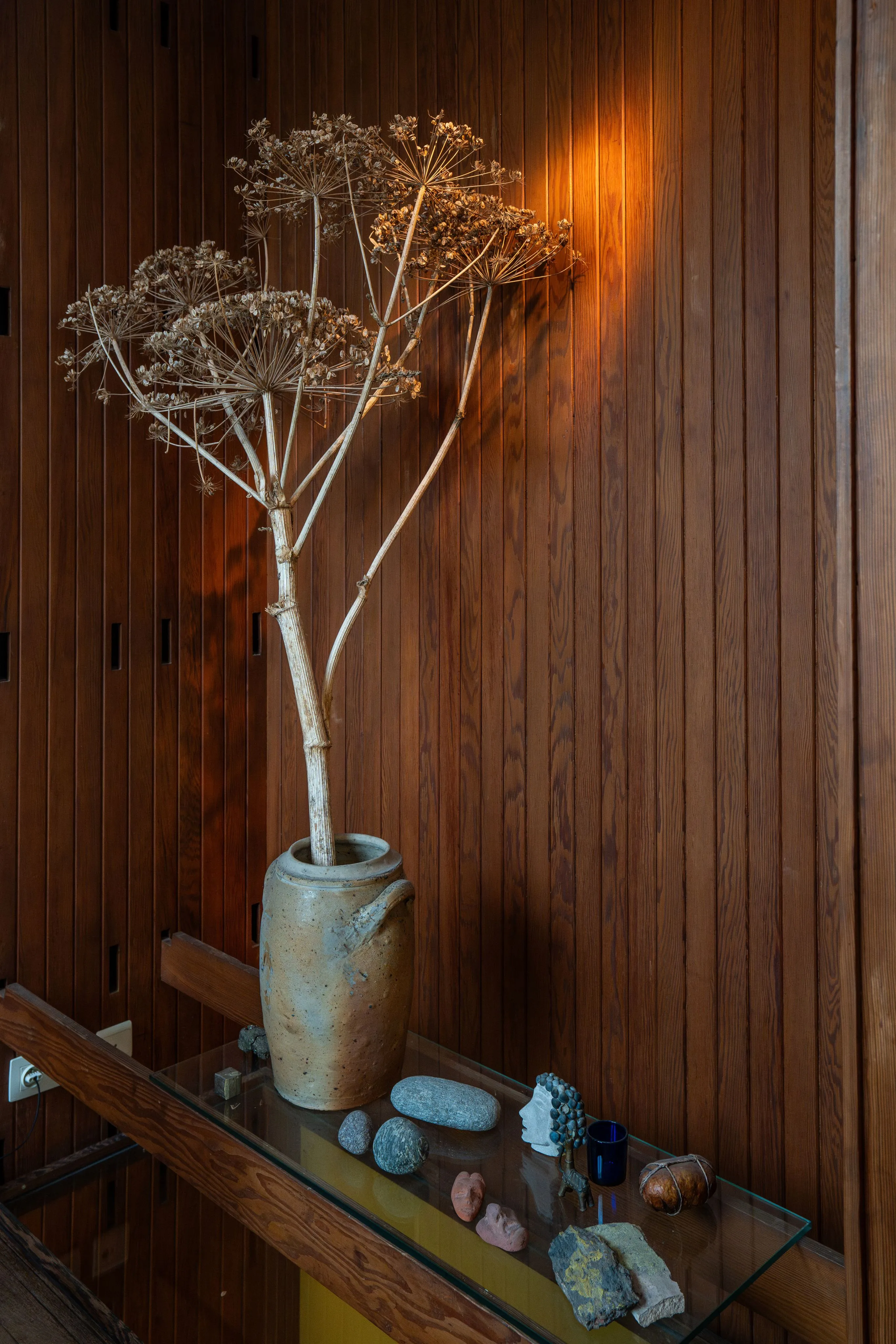 Wood-paneled interior with a rustic ceramic vase holding tall dried plant stems on a glass-topped console table, surrounded by small stones and decorative objects under warm lighting.