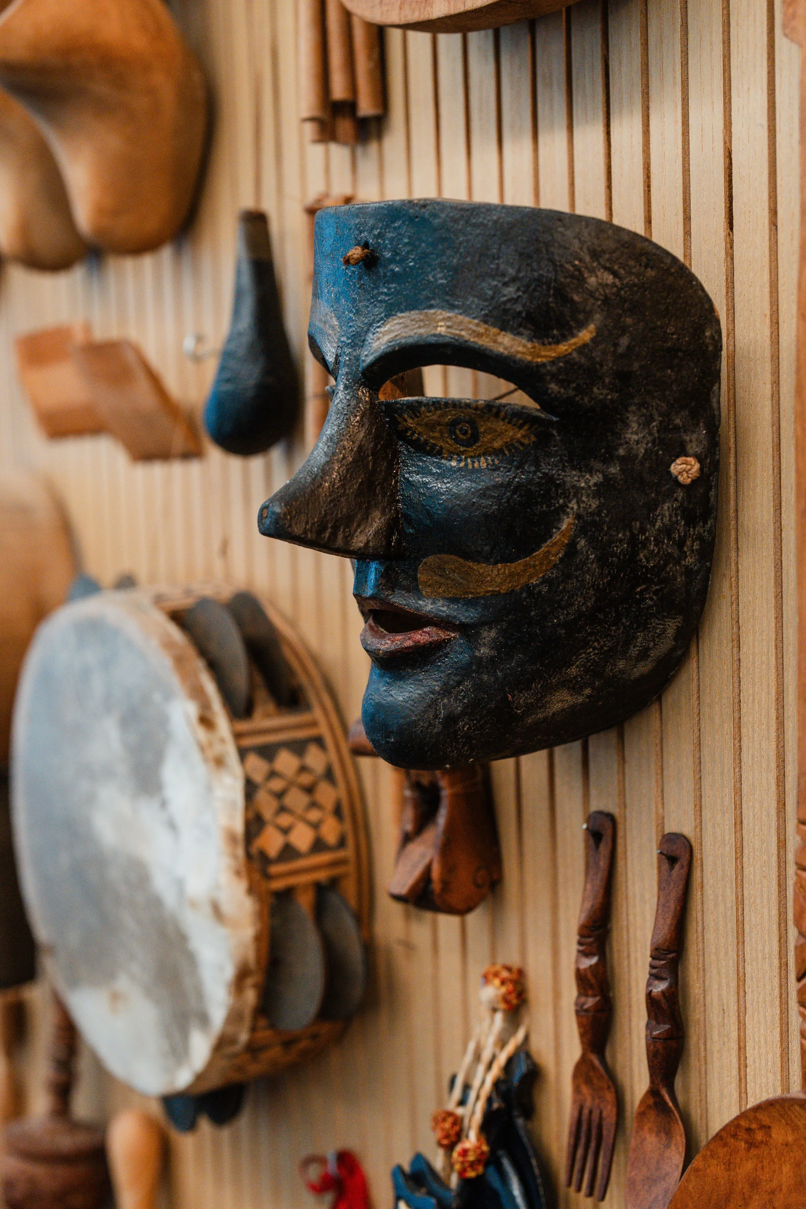 A close-up of a wall display featuring a large dark blue and black mask, a hand drum, and various wooden utensils and artifacts.