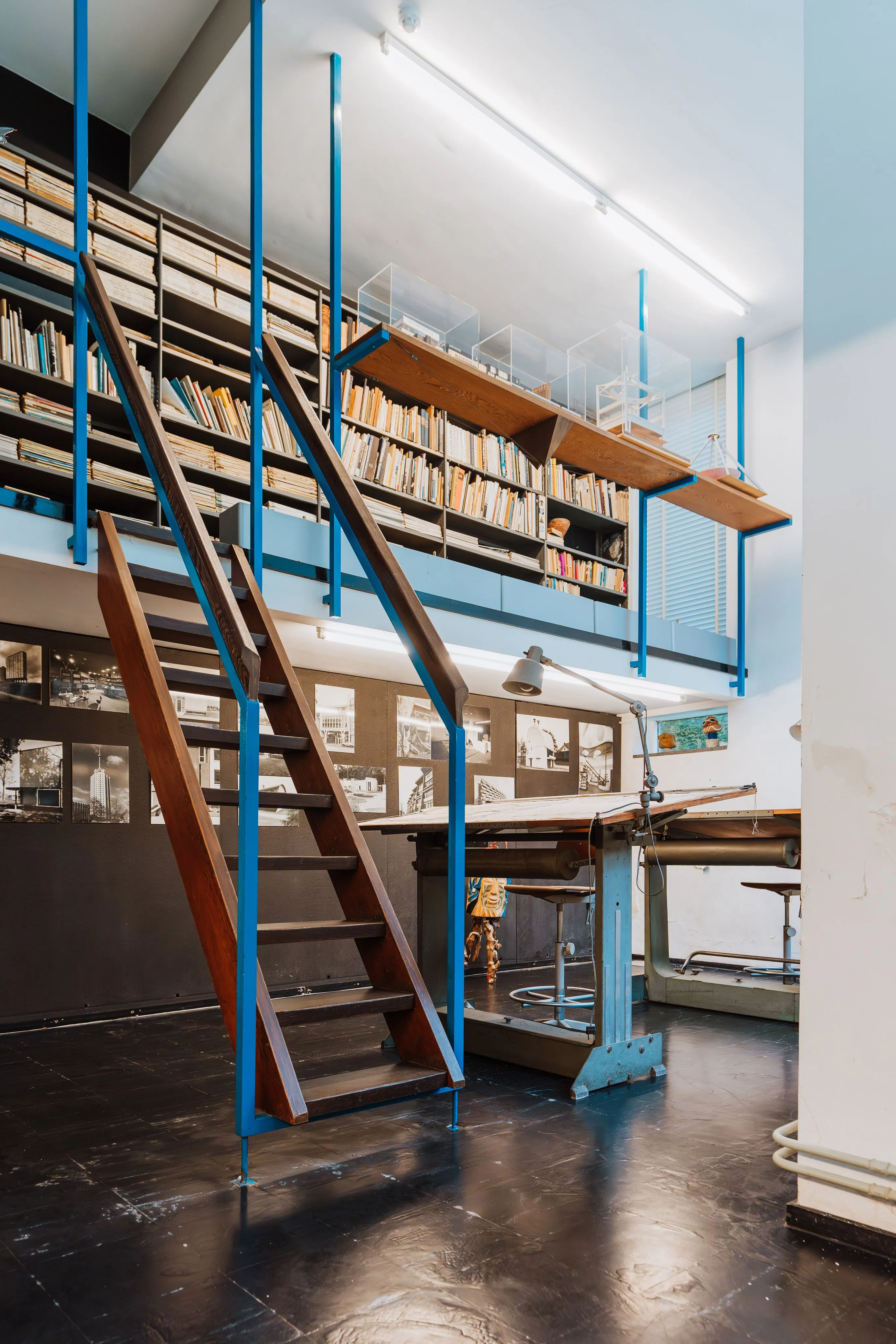 Escalera de madera con estructura metálica azul que asciende a un entresuelo lleno de estanterías, con una mesa de dibujo y planos arquitectónicos visibles abajo en el interior de un estudio modernista.