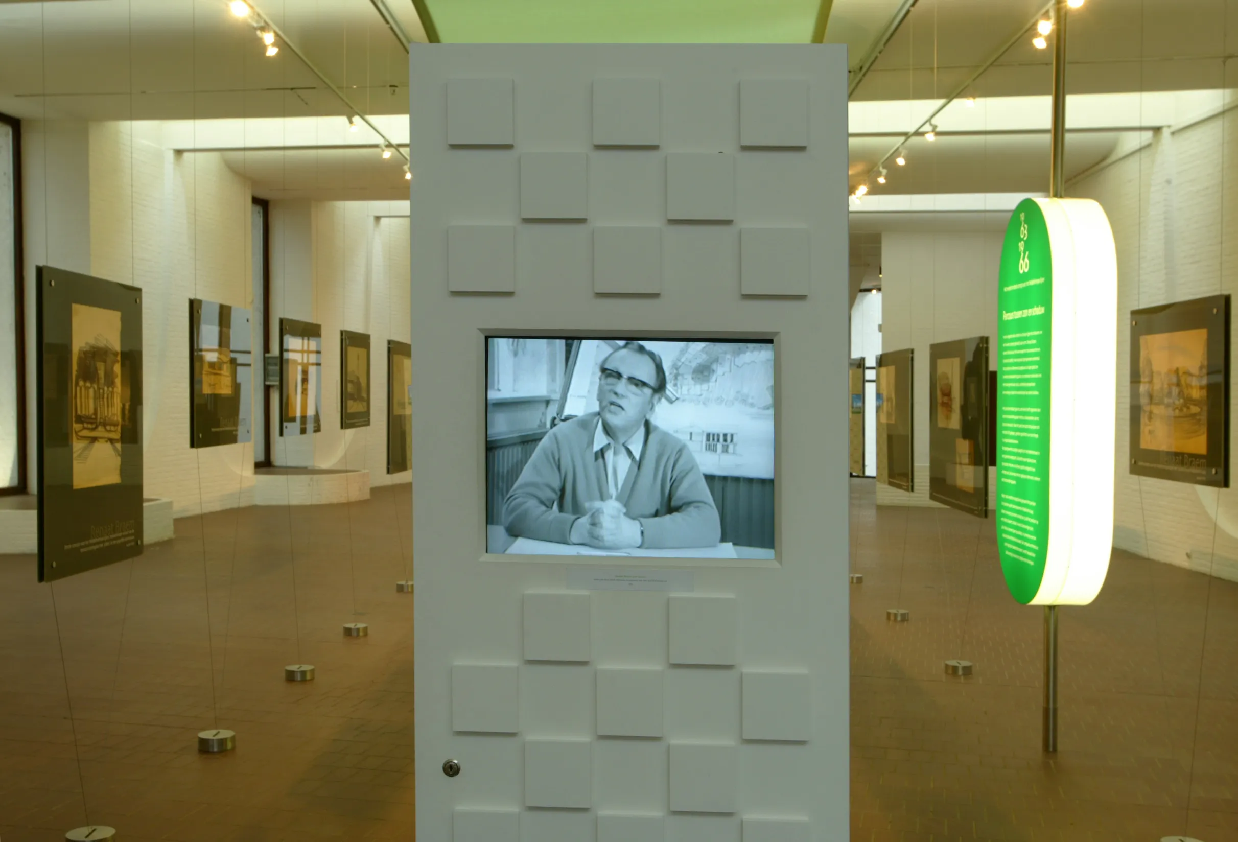 Espacio de exposición interior con una columna blanca texturizada que presenta una fotografía en blanco y negro de un hombre incrustada, obras de arte enmarcadas en las paredes, iluminación de riel y un letrero informativo ovalado de color verde sobre un poste.