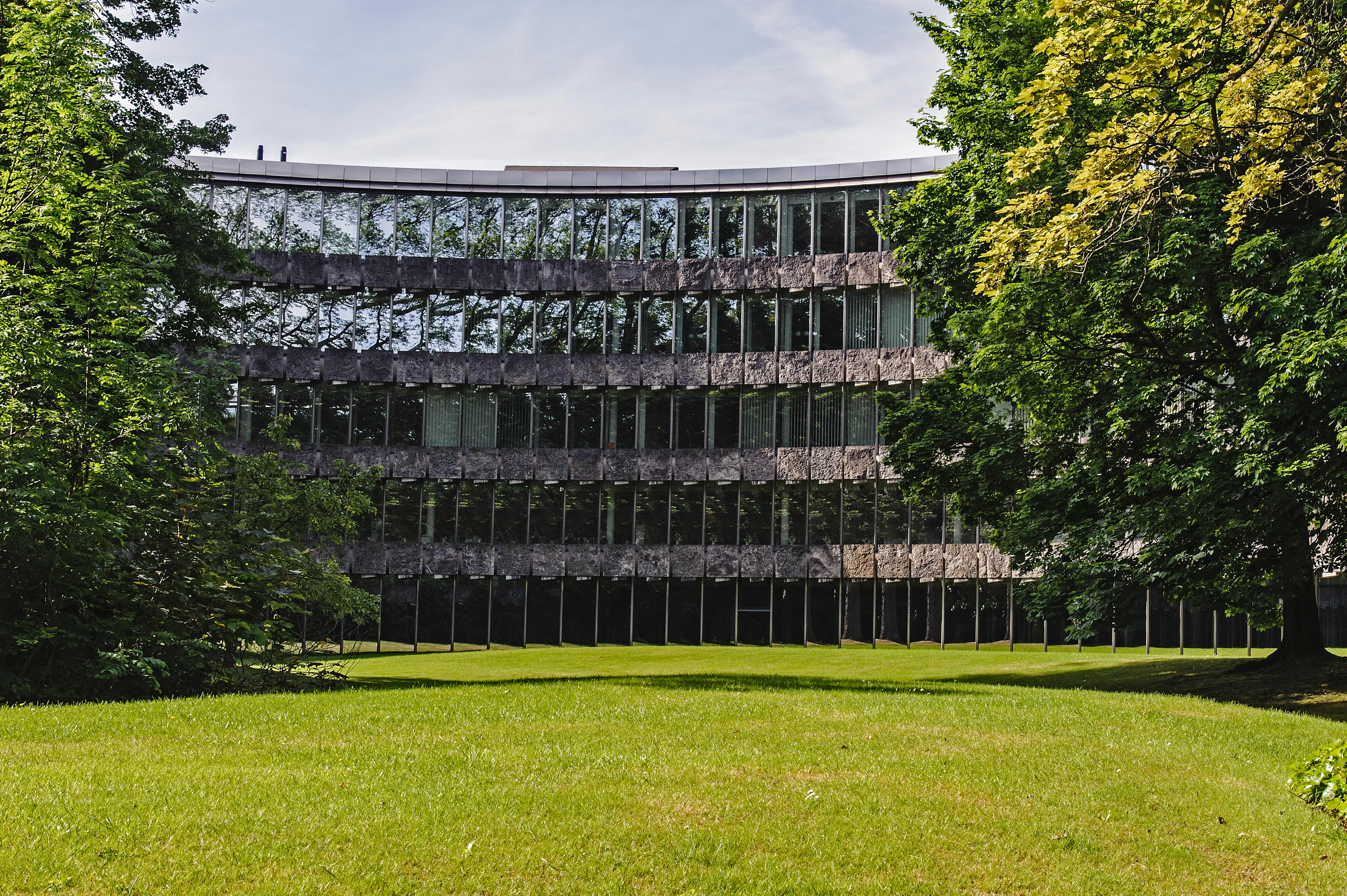 Edificio modernista curvo de varias plantas con franjas horizontales de ventanales, enmarcado por árboles verdes y un césped en primer plano.