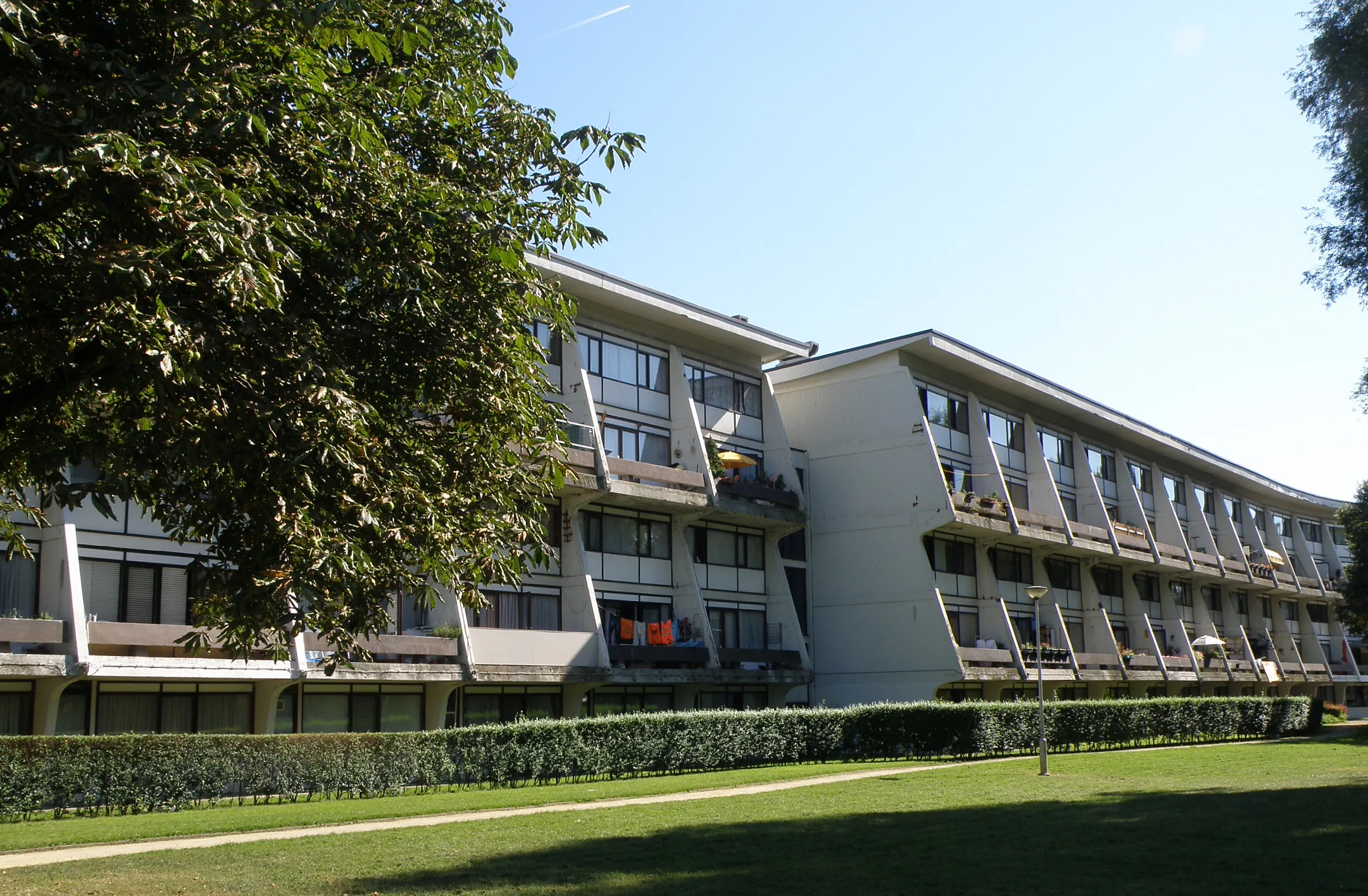 Largo edificio de apartamentos modernista de color blanco con balcones repetitivos y un césped verde en la parte delantera, enmarcado por árboles bajo un cielo azul.