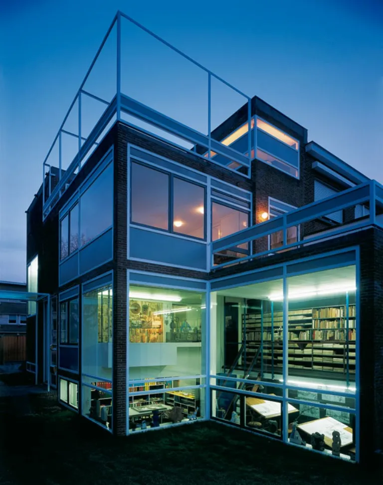 Vista exterior de la Casa Renaat Braem al atardecer, que presenta una estructura modernista de ladrillo y cristal con habitaciones interiores iluminadas y una terraza en la azotea.