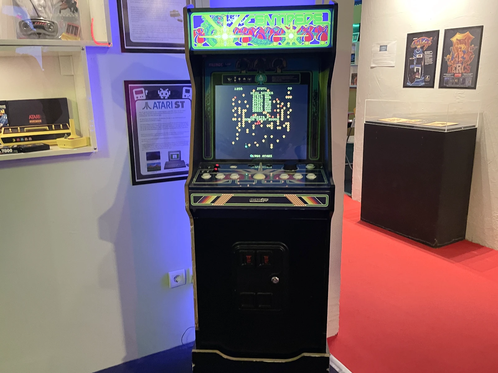 Een vintage arcadekast met een gloeiend scherm waarop een gepixelde game wordt weergegeven, in een museum met rood tapijt en vitrines.
