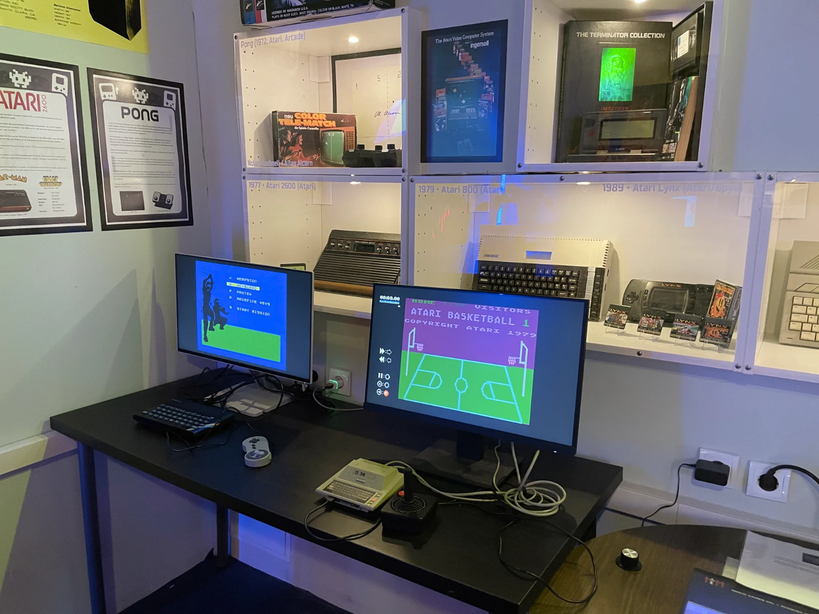 Dos monitores de computadora que muestran juegos retro en un escritorio con equipo de juego vintage y estantes de máquinas arcade y consolas al fondo