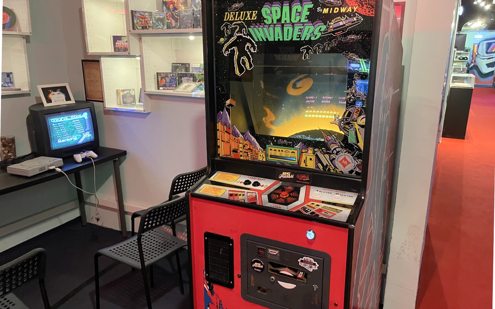 Máquina arcade 'Space Invaders' roja con pantalla e panel de control iluminados, rodeada de estantes con videojuegos y una configuración de ordenador retro.