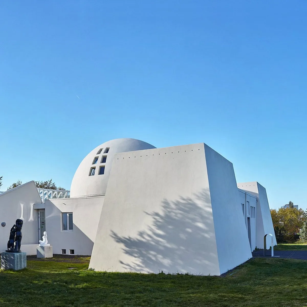 Edificio geométrico blanco con cúpula y paredes angulares, rodeado por un césped verde con esculturas, bajo un cielo azul despejado