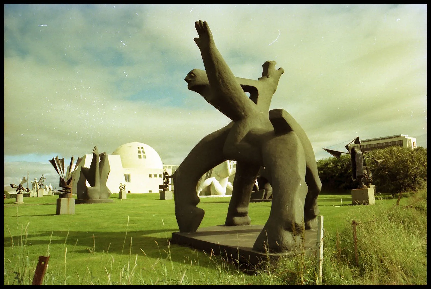 Jardín de esculturas al aire libre con grandes estatuas de bronce sobre un césped verde, con un edificio blanco en forma de cúpula al fondo bajo un cielo nublado.