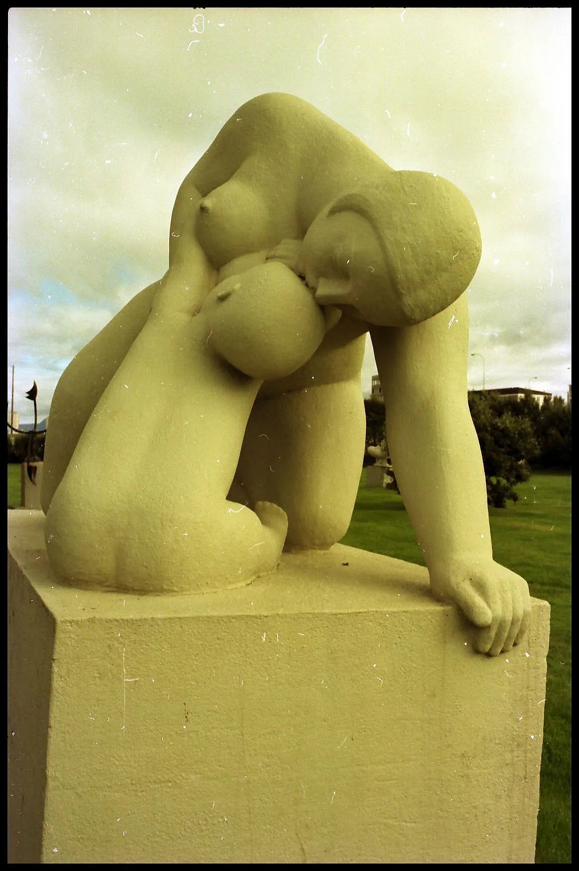 Stenen sculptuur van een naakte vrouwelijke figuur die voorover leunt op een rechthoekig voetstuk in een tuin.