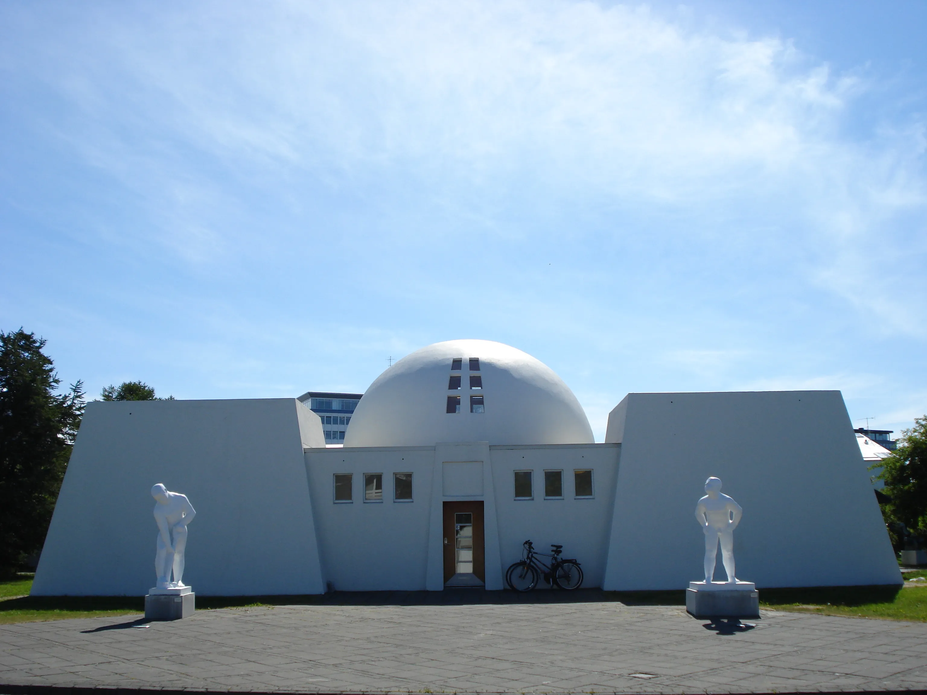 Edificio blanco con cúpula y dos esculturas blancas flanqueando la entrada, bicicletas aparcadas cerca de la puerta bajo un cielo azul