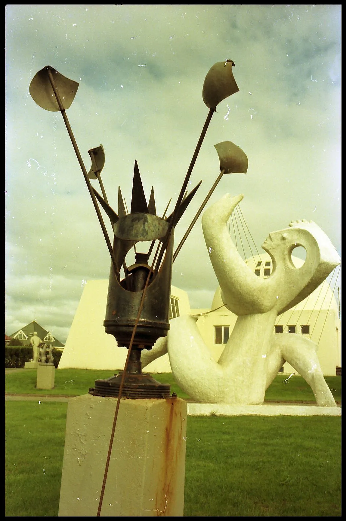 Jardín de esculturas que presenta una escultura de metal oscuro con tallos altos sobre un pedestal y una gran figura abstracta blanca, sobre el fondo del edificio del museo y un césped verde.