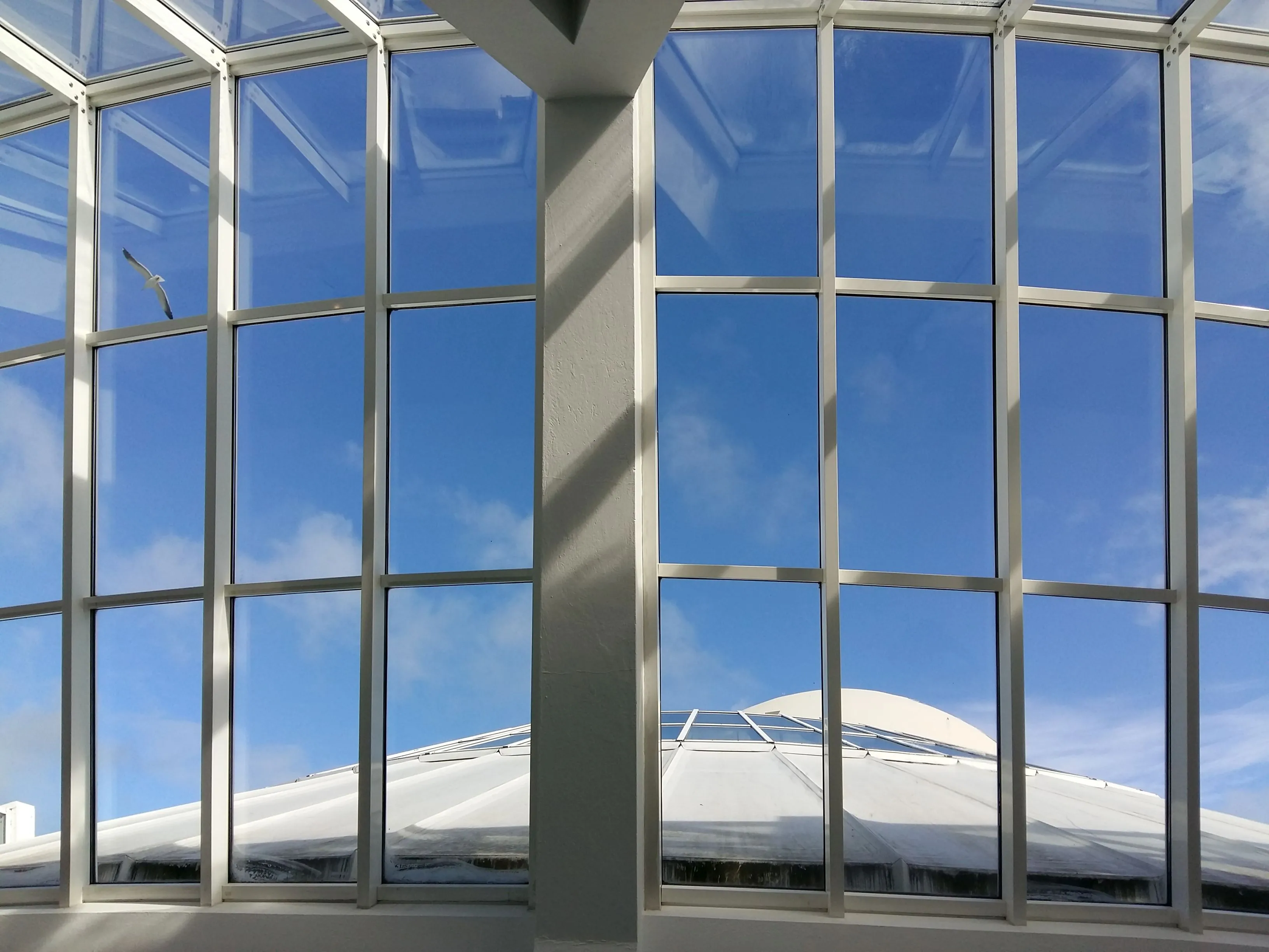 Gran muro cortina de cristal con marcos de metal y una columna central dentro de un edificio, con vistas a una estructura en forma de cúpula blanca sobre un cielo azul con nubes