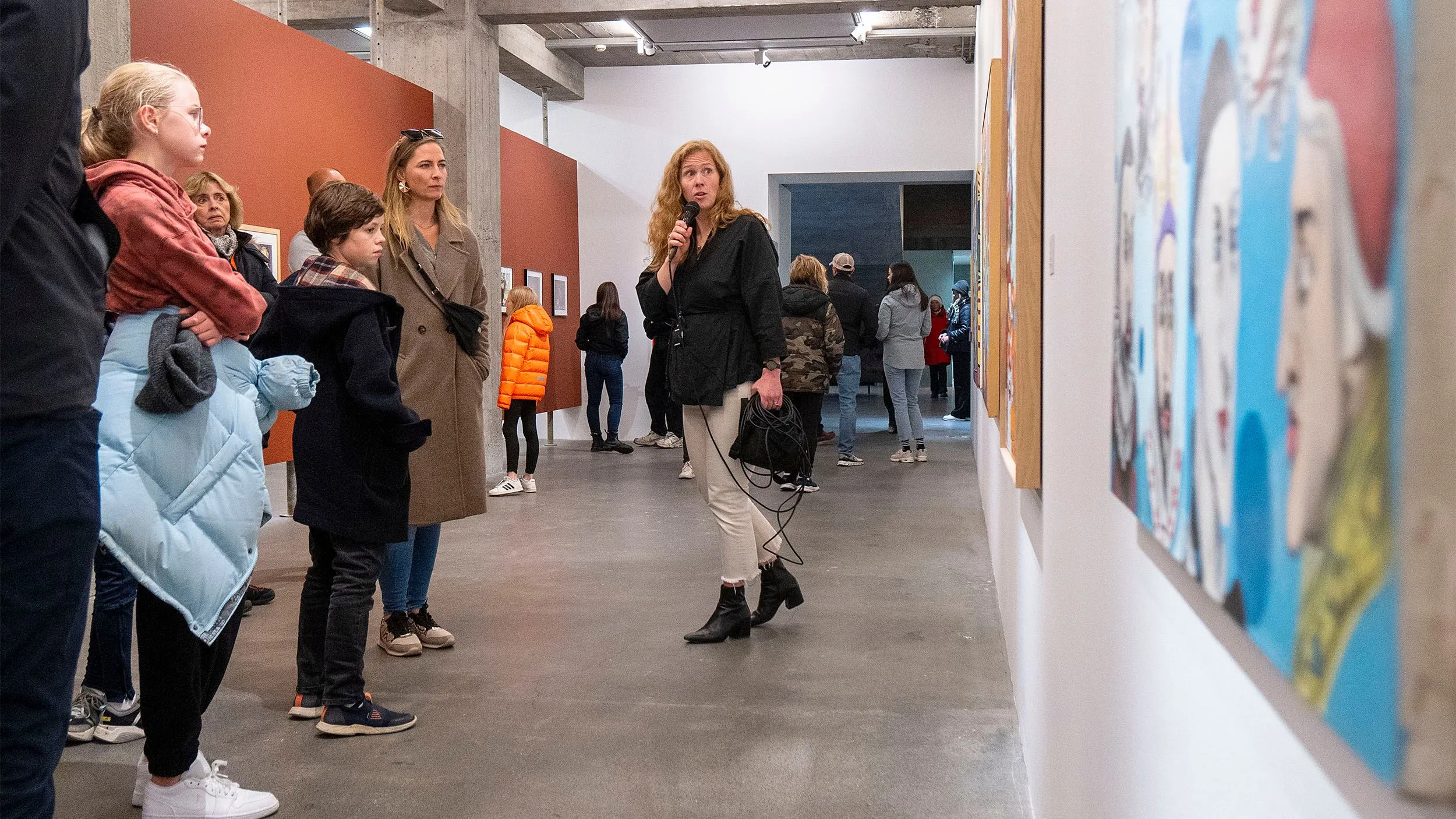 Un grupo de visita guiada compuesto por adultos y niños se encuentra en la sala de un museo escuchando a un guía que habla por un micrófono, con cuadros coloridos visibles en la pared derecha.