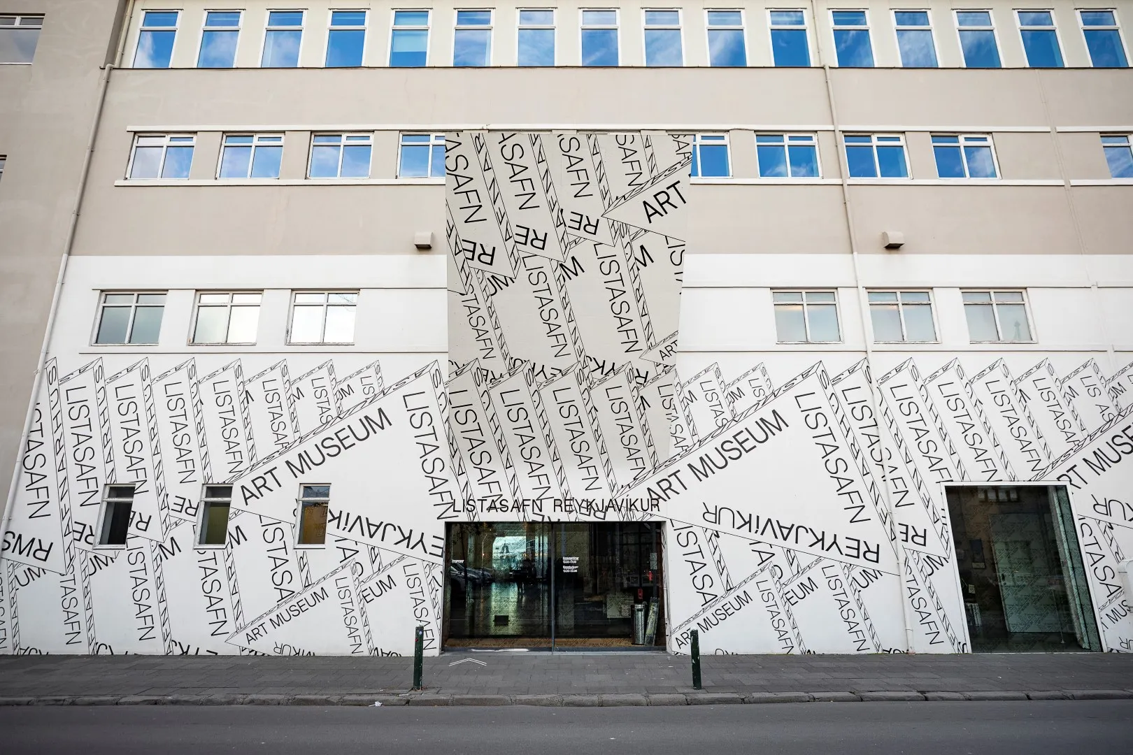 Fachada blanca del edificio cubierta con el texto repetido 'Reykjavík Art Museum', con puertas de entrada de cristal y filas de ventanas tintadas de azul encima.