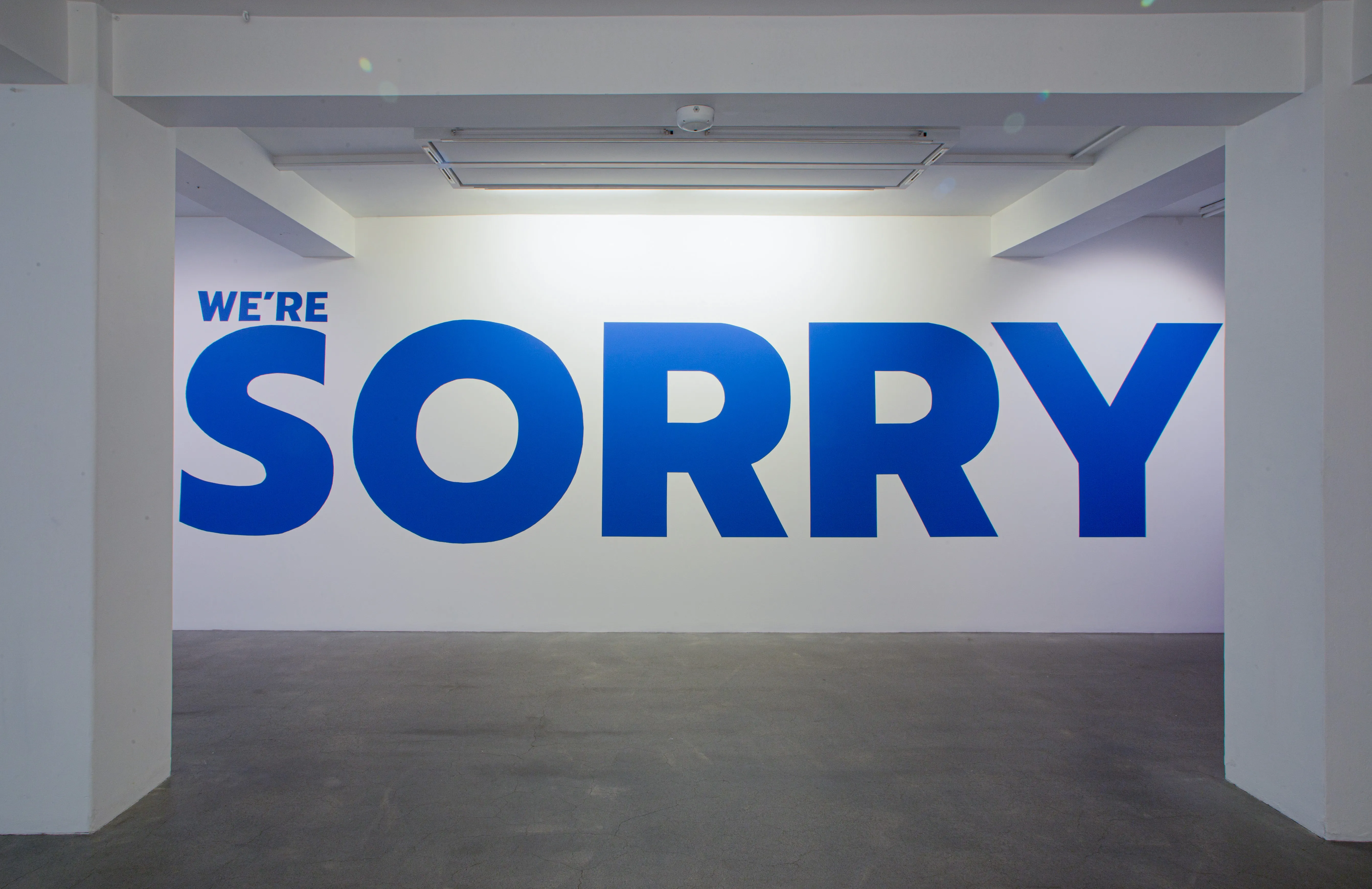 Gran mural azul con la inscripción "WE'RE SORRY" pintado en una pared blanca de una galería, visto a través de una puerta abierta en un espacio de museo.