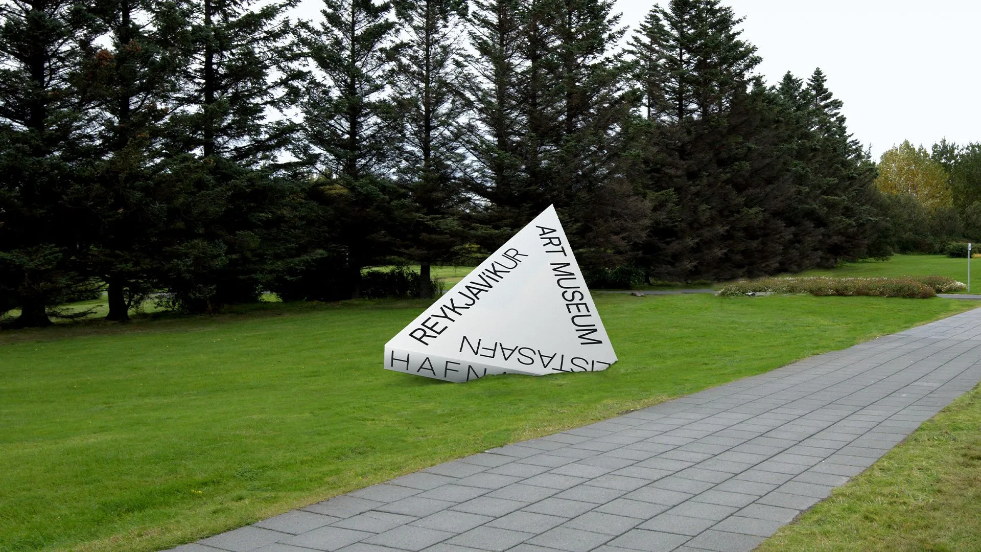 Gran escultura triangular blanca con el letrero 'Reykjavíkur Listasafn Hafnar / Art Museum' sobre un césped verde con pinos y un camino pavimentado