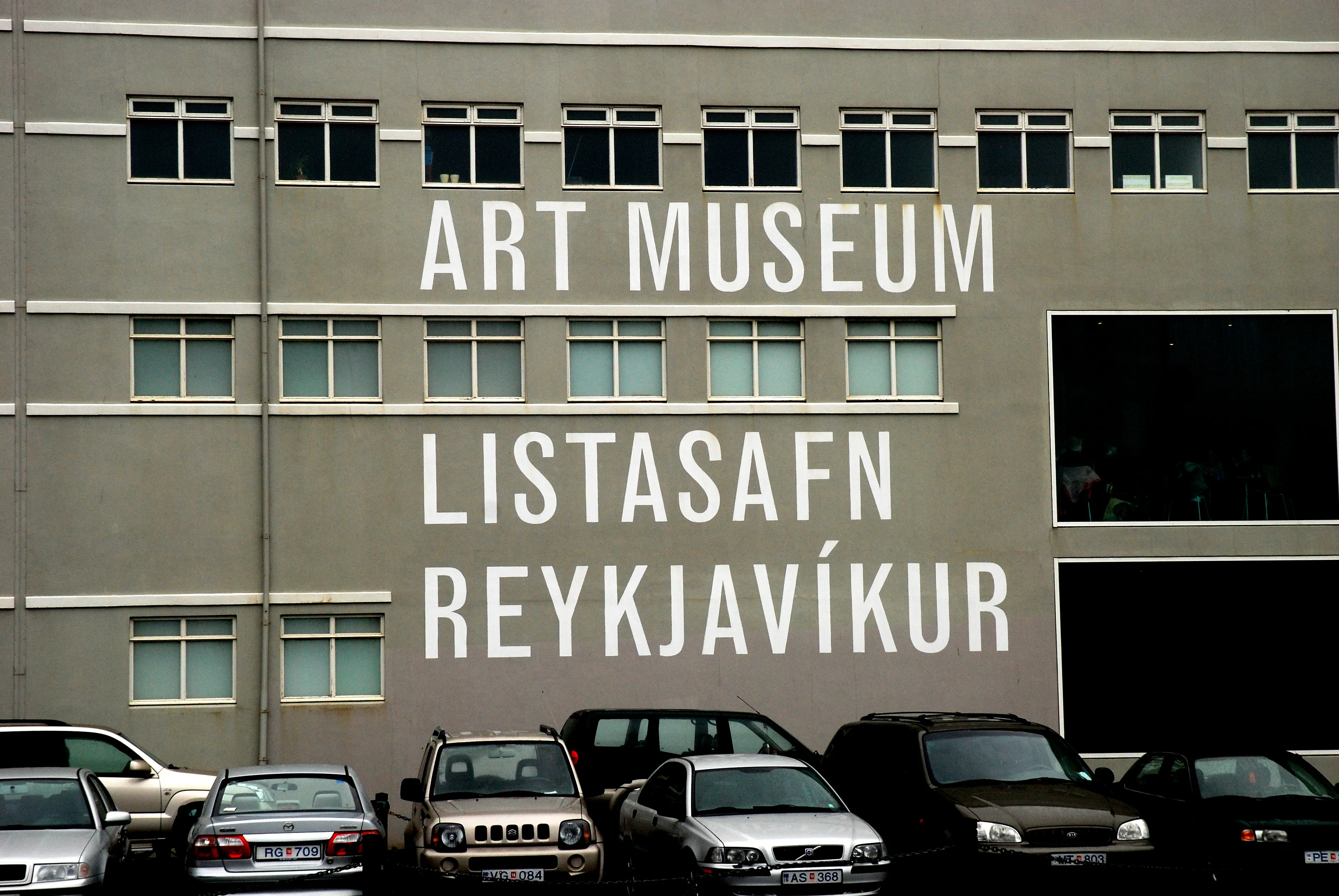 Szara fasada budynku z dużym napisem malowanym na biało o treści 'ART MUSEUM' oraz 'LISTASAFN REYKJAVÍKUR', z rzędem zaparkowanych samochodów u podnóża.