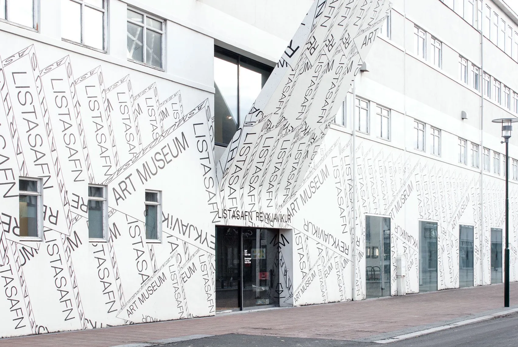 Fachada de edificio blanco cubierta con un revestimiento tipográfico a gran escala que repite las palabras LISTASAFN REYKJAVÍKUR ART MUSEUM en texto negro, con una entrada de cristal visible a nivel de calle.