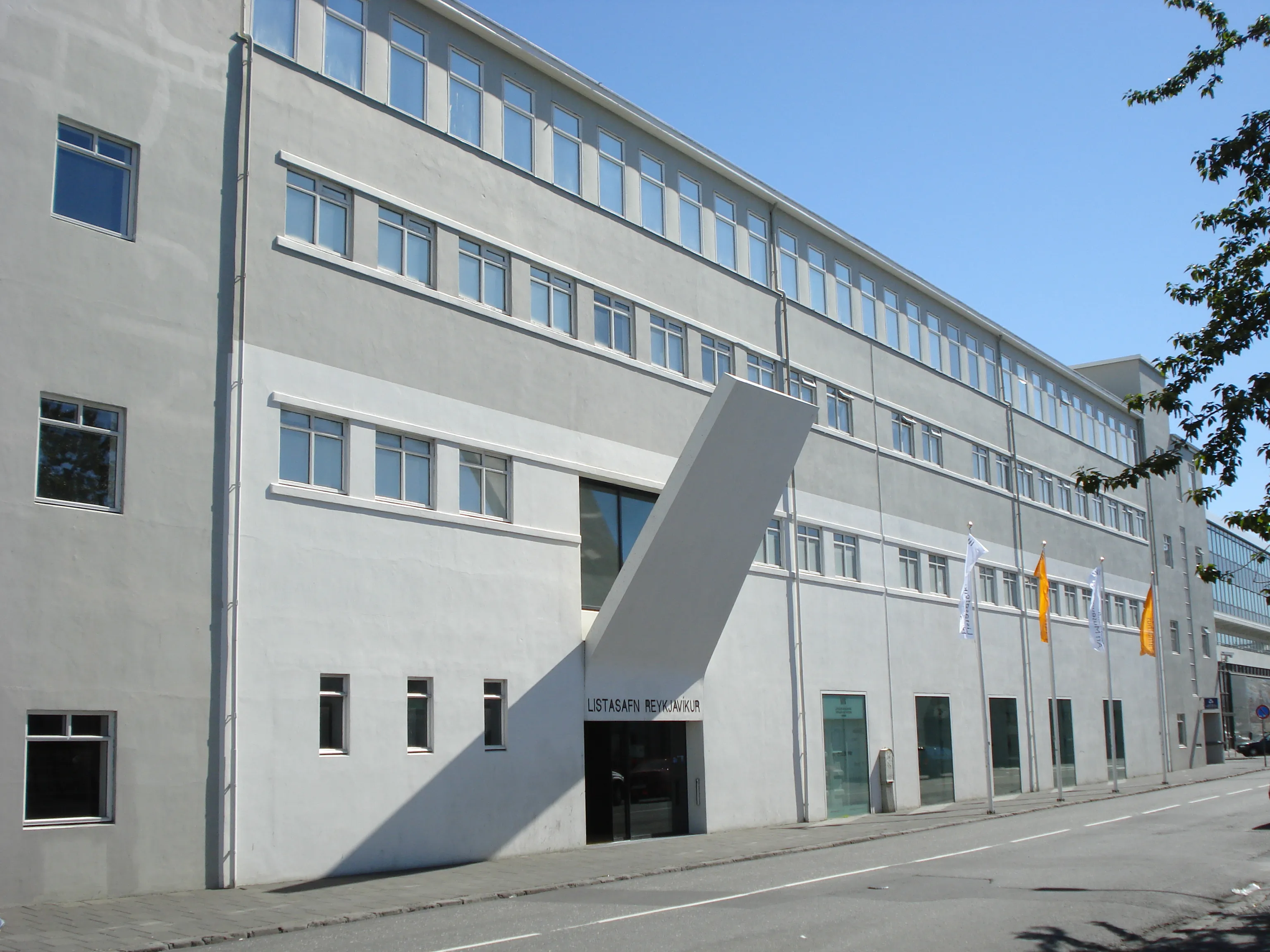 Fasada Reykjavík Art Museum, dużego, jasnoszarego modernistycznego budynku z charakterystycznym ukośnym zadaszeniem nad wejściem i flagami powieszonymi na elewacji.