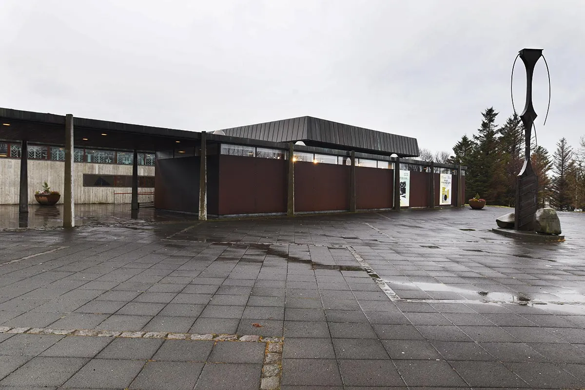 Widok z zewnątrz na budynek muzeum Kjarvalsstaðir z dużą, ciemną, abstrakcyjną rzeźbą na mokrym, wybrukowanym placu pod zachmurzonym niebem.