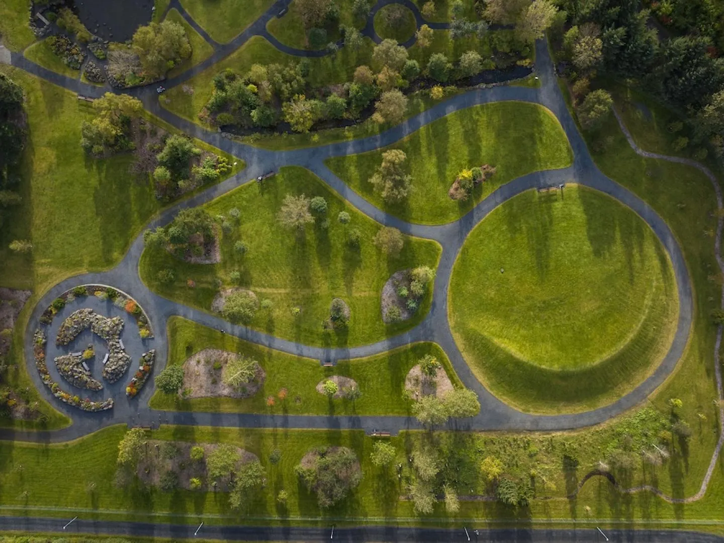 Luchtfoto van kronkelende grijze paden door groene gazons en boomgroepen in een botanische tuin, met een cirkelvormig stenen arrangement aan de linkerzijde en een groot rond gazon aan de rechterzijde.