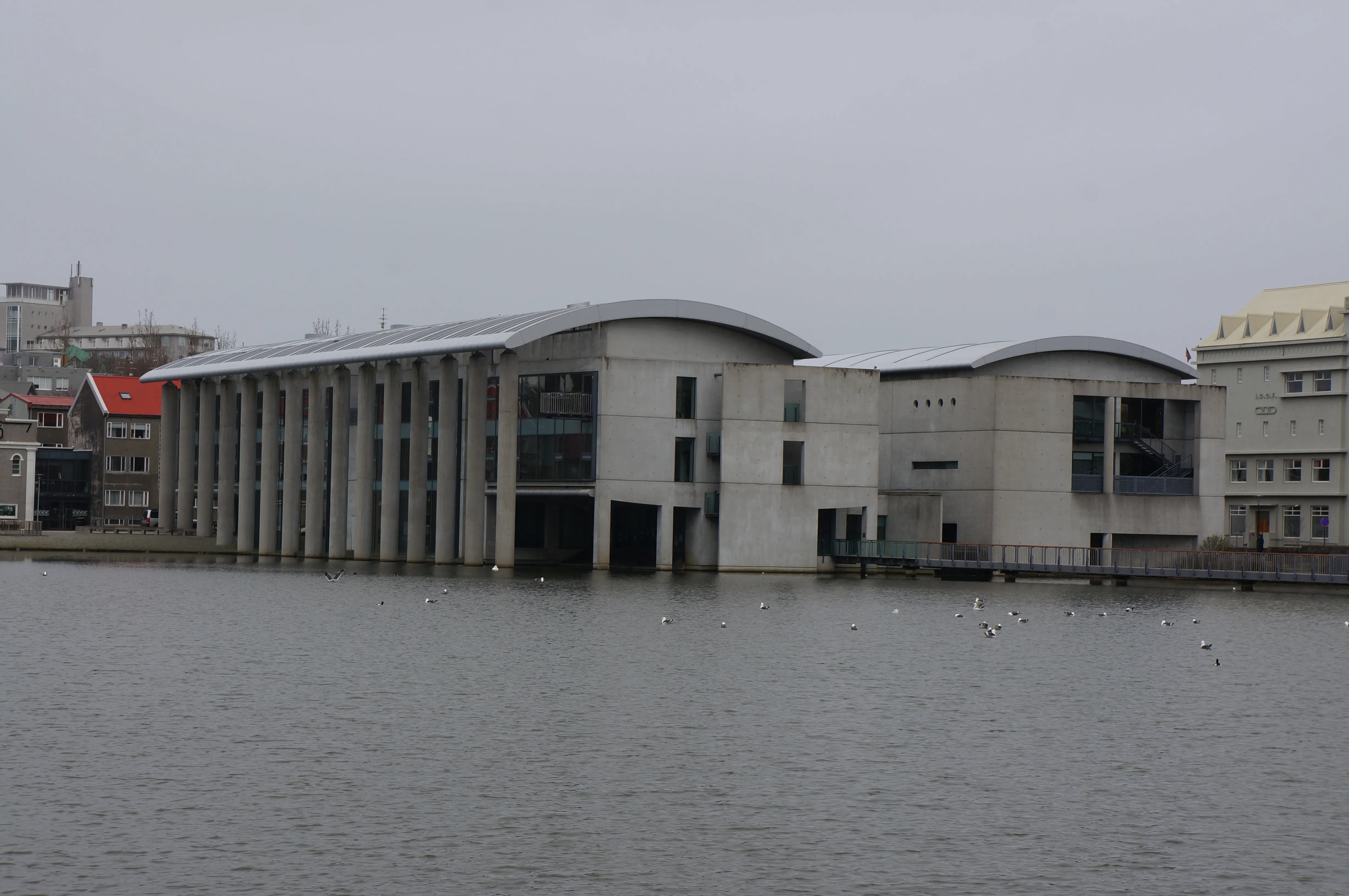Edificio moderno de hormigón con techo curvo y columnas que se extienden sobre un lago, visto desde el otro lado del agua en un día nublado