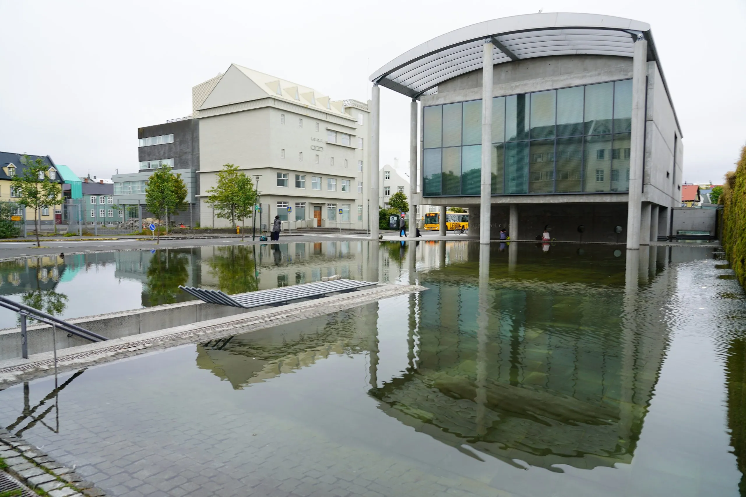 Moderno edificio del Ayuntamiento de Reykjavík con fachada de cristal reflejado en un estanque poco profundo, con una pasarela peatonal y cielo nublado.