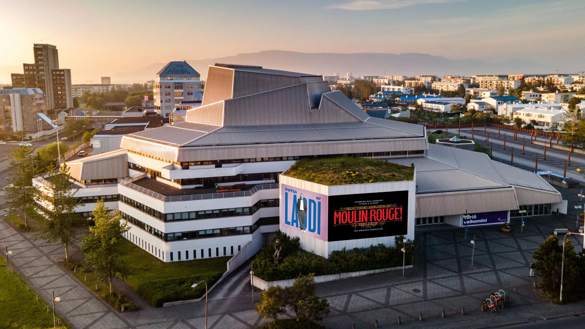 Vista aérea del edificio del Reykjavík City Theatre al atardecer con un anuncio de Moulin Rouge en su fachada, rodeado por edificios de la ciudad y montañas al fondo.