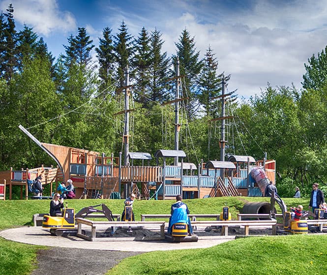 Plac zabaw na świeżym powietrzu w Reykjavík Family Park and Zoo z dużą drewnianą konstrukcją do wspinaczki w kształcie statku, jeździkami oraz odwiedzającymi na trawiastym terenie otoczonym sosnami pod błękitnym niebem.