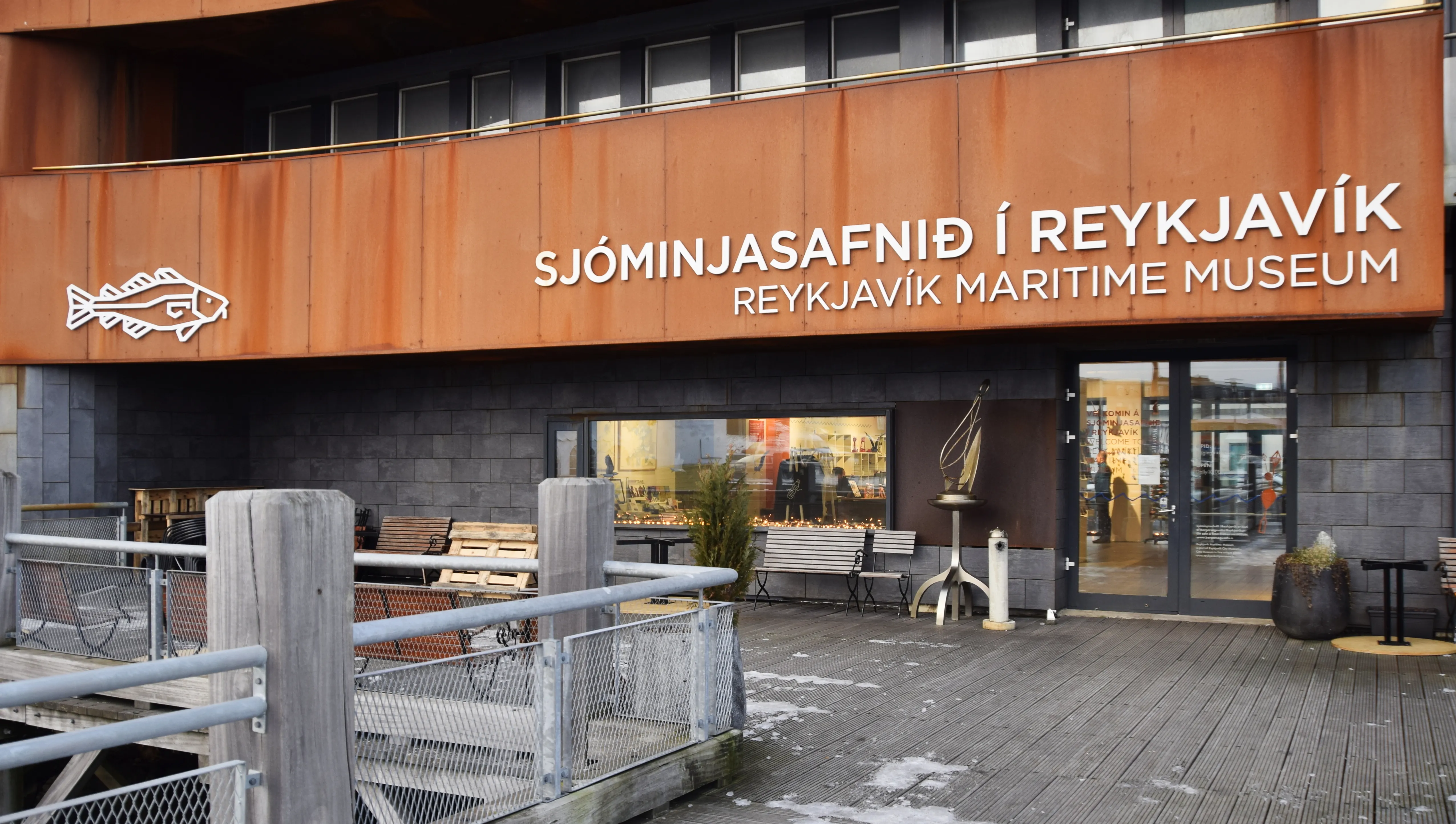 Exterior facade of the Reykjavík Maritime Museum showing rust-colored wooden cladding with white bilingual signage and a fish skeleton logo above a wooden deck entrance.