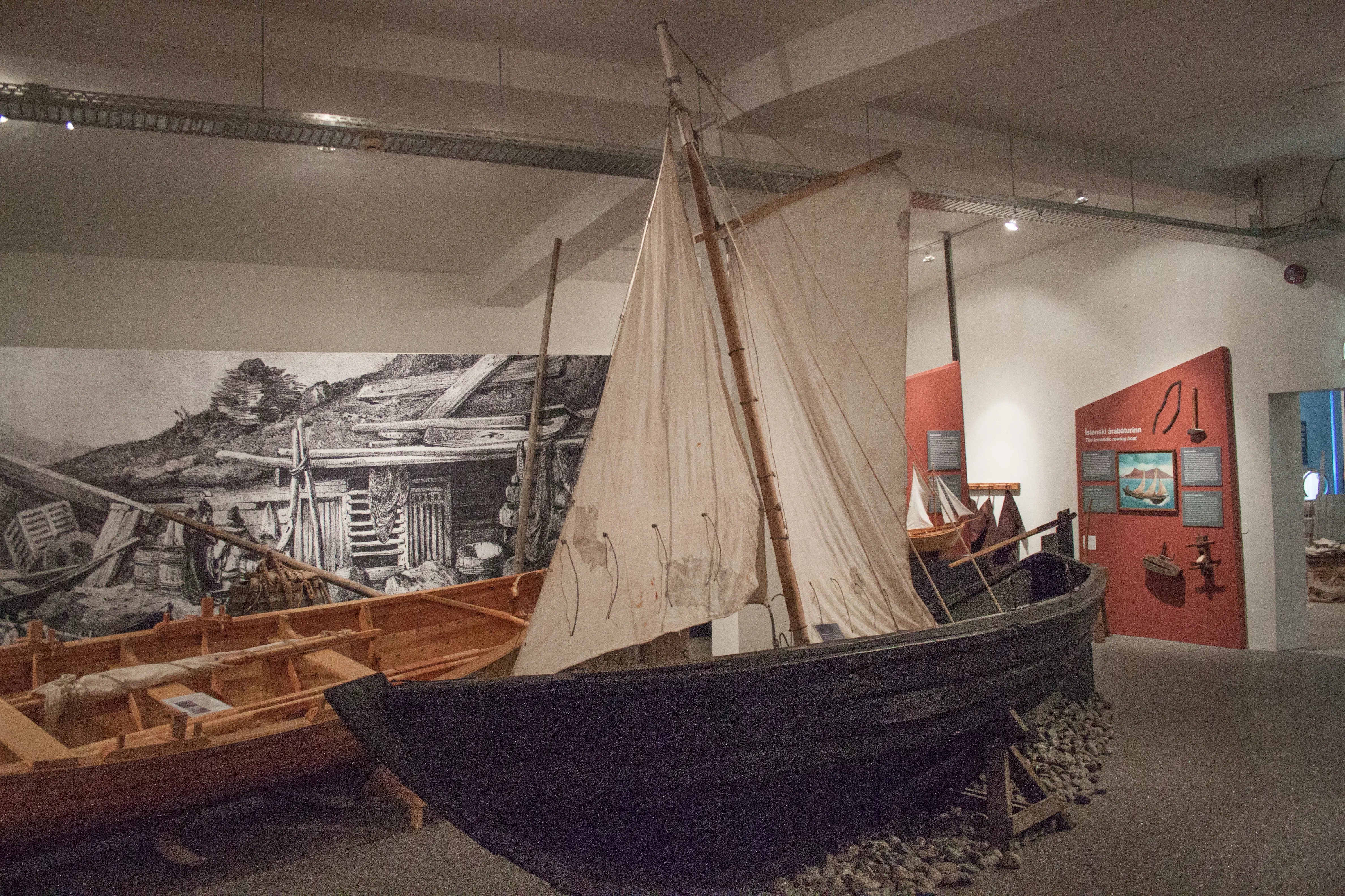 A traditional Icelandic fishing boat with a white sail displayed indoors on a bed of stones, with a large black-and-white harbor mural on the wall behind it.