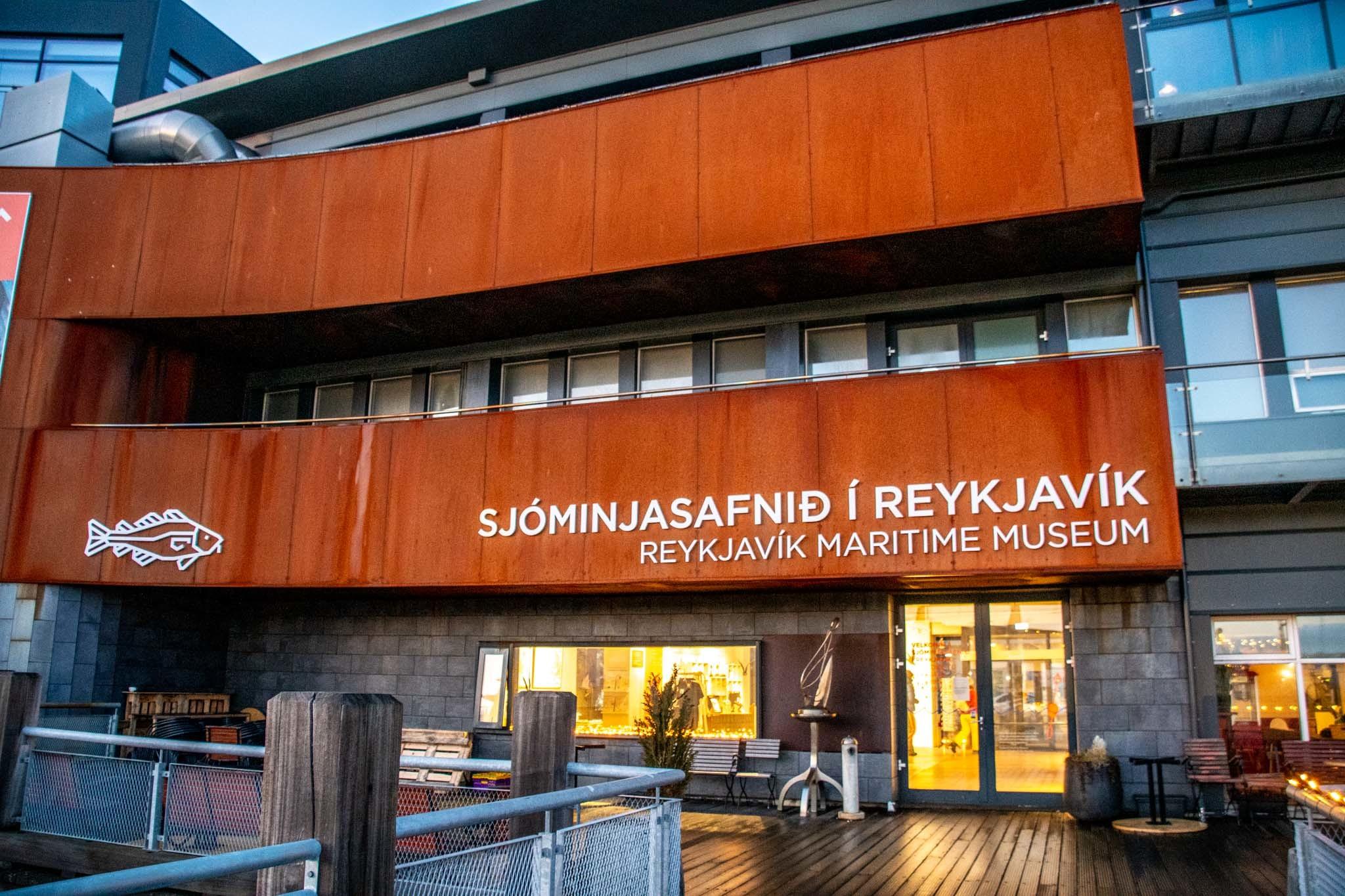 Rust-colored corten steel facade of the Reykjavík Maritime Museum with bilingual signage and a white fish logo, showing the glass entrance doors with warm interior lighting and a wooden deck terrace in front