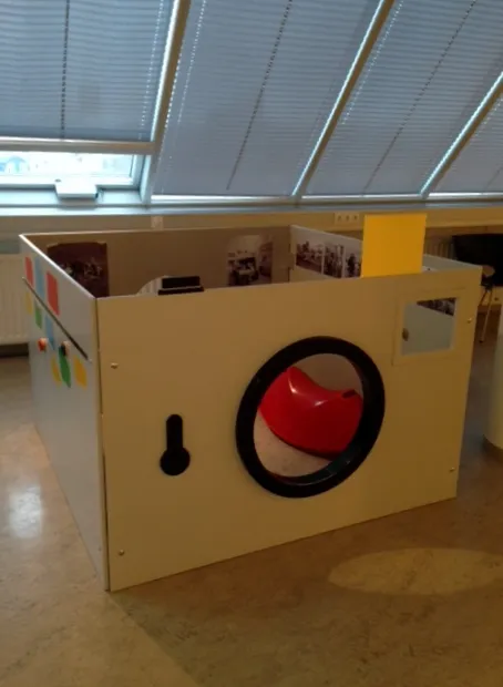 Una gran estructura de juego de madera con forma de cámara, con un asiento rojo dentro de la abertura circular del objetivo, expuesta en una sala de museo con tragaluces y fotografías en las paredes interiores.