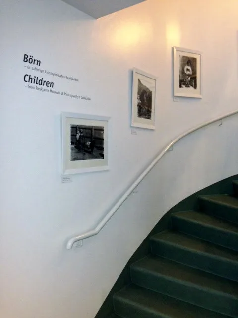 Pared interior del Reykjavík Museum of Photography que muestra la exposición 'Börn / Children' con tres fotografías en blanco y negro enmarcadas sobre una escalera curva de color verde con un pasamanos blanco.