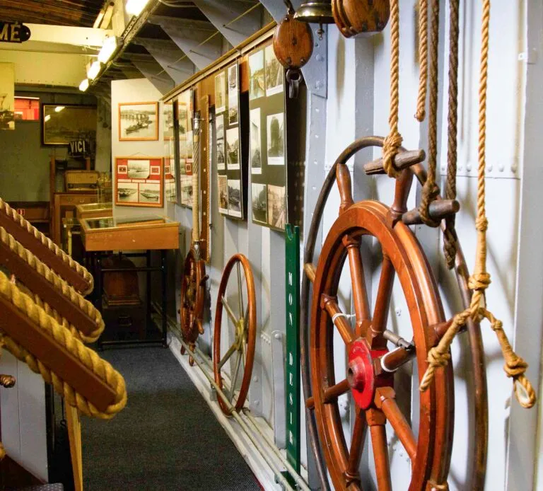 Interior de un museo naval con timones de madera, cuerdas gruesas y paneles informativos a lo largo de un pasillo estrecho.
