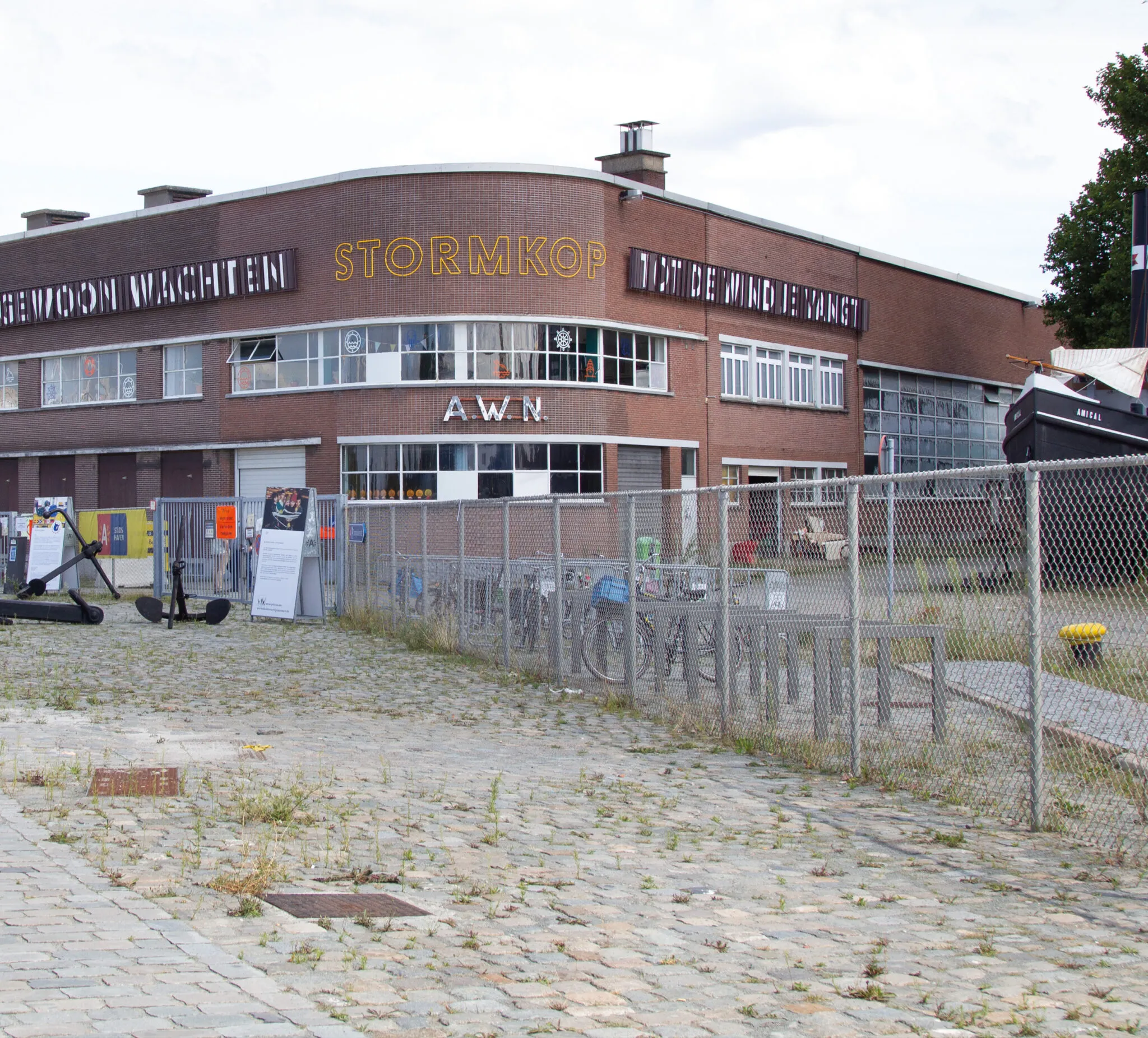 Exterior de un edificio de ladrillo marrón curvo con el letrero 'STORMKOP', rodeado por una valla de malla metálica en un terreno adoquinado.