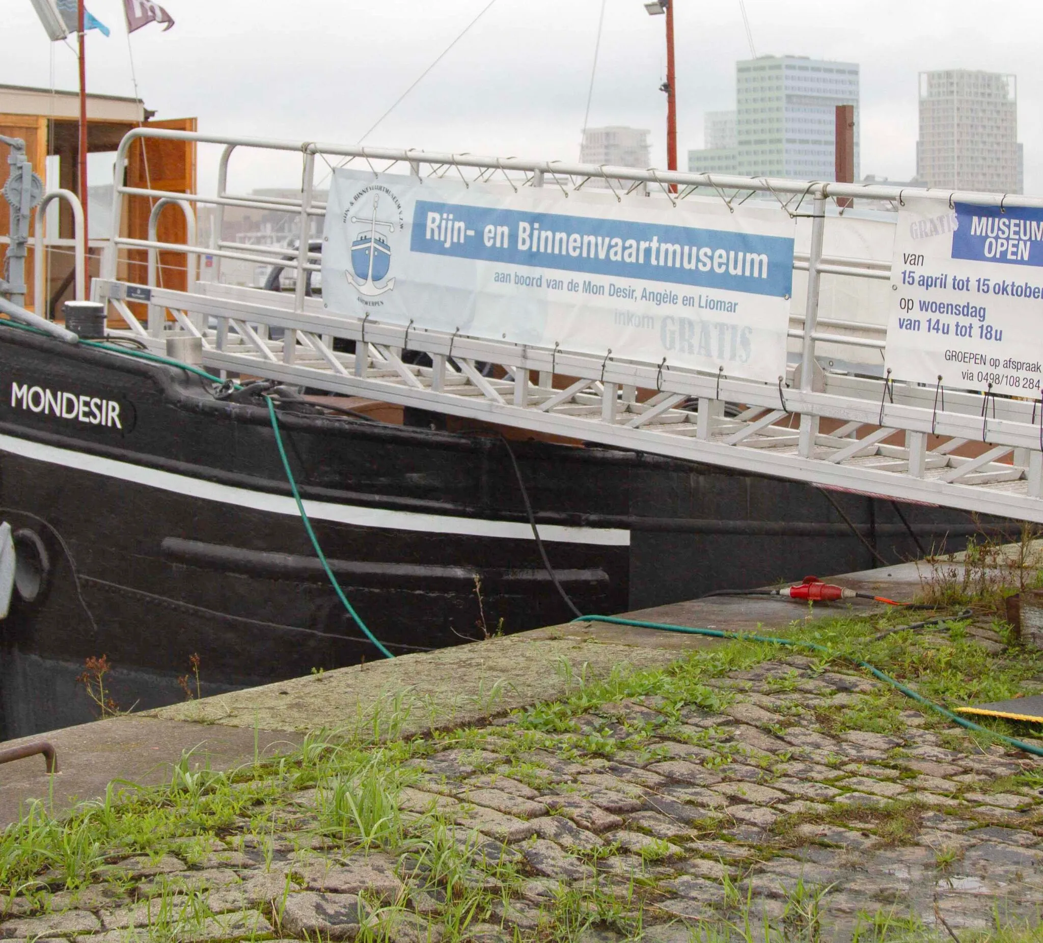 Casco de barco negro llamado Mondesir atracado en un muelle de adoquines, con una pasarela blanca que muestra un cartel para el Rijn- en Binnenvaartmuseum.