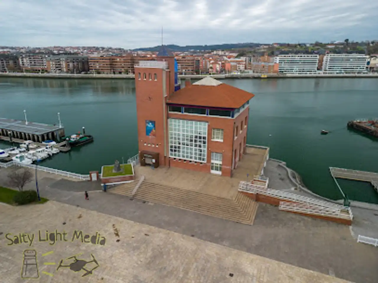 Vista aérea de un edificio de museo de ladrillo rojo con una torre alta y grandes ventanales de cristal a la orilla del agua, con un puerto deportivo y el horizonte de la ciudad al otro lado del río