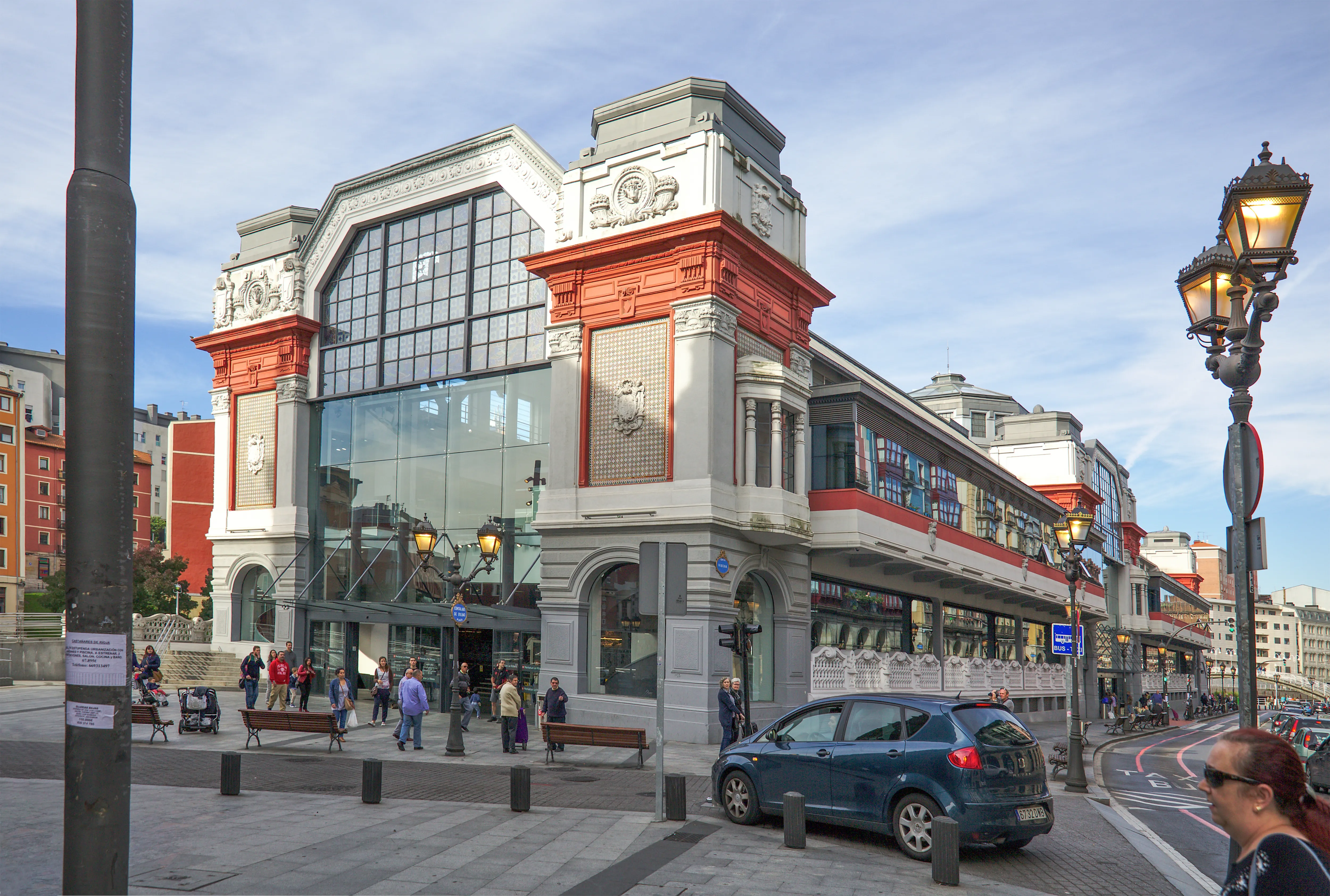 Vista exterior del Mercado de la Ribera en Bilbao, un gran edificio de estilo art déco con fachada roja y gris, grandes ventanales de cristal y detalles en piedra, con peatones y un coche en la calle frente a él.
