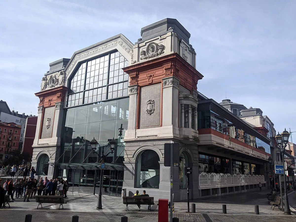 Vista exterior del Mercado de la Ribera en Bilbao, que presenta arquitectura Art Déco con una gran fachada de cristal, ornamentación en piedra y detalles en ladrillo rojo.