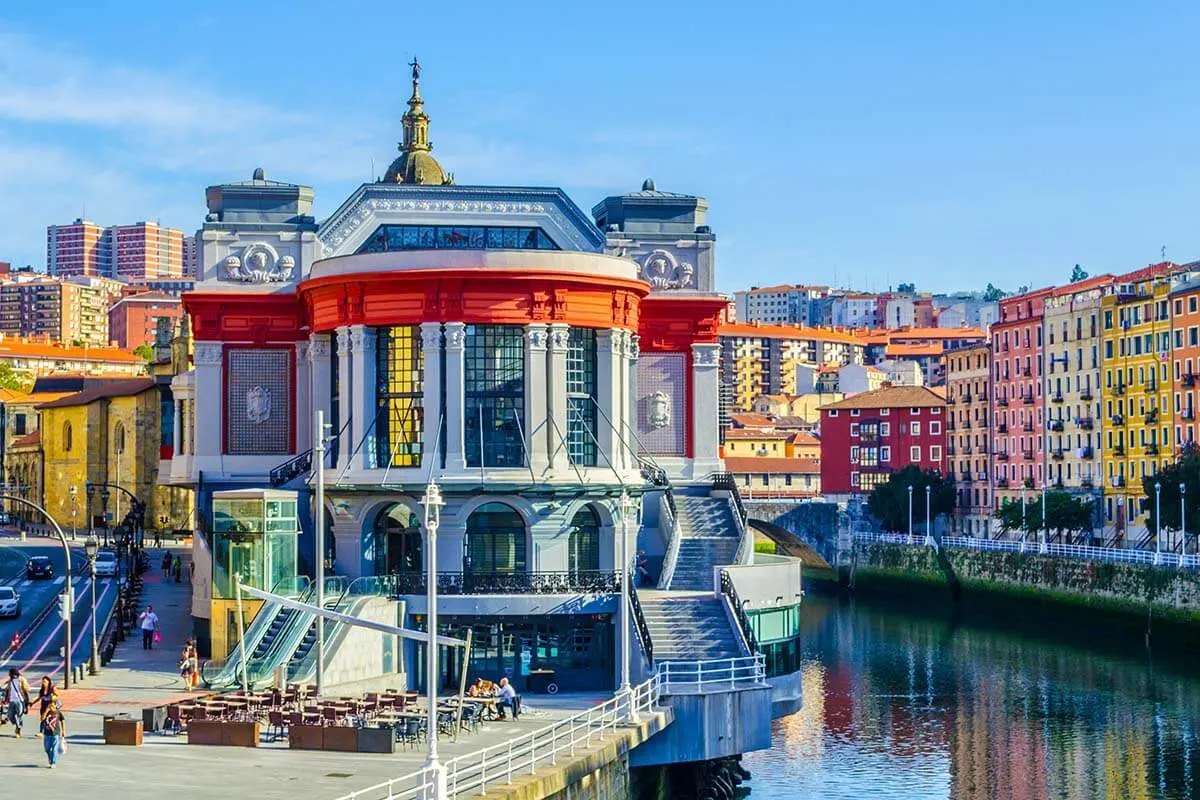 Vista exterior del Mercado de la Ribera en Bilbao, un edificio art déco con detalles en rojo y una torre ornamentada situado junto a la ría, con los coloridos edificios de la ciudad al fondo.