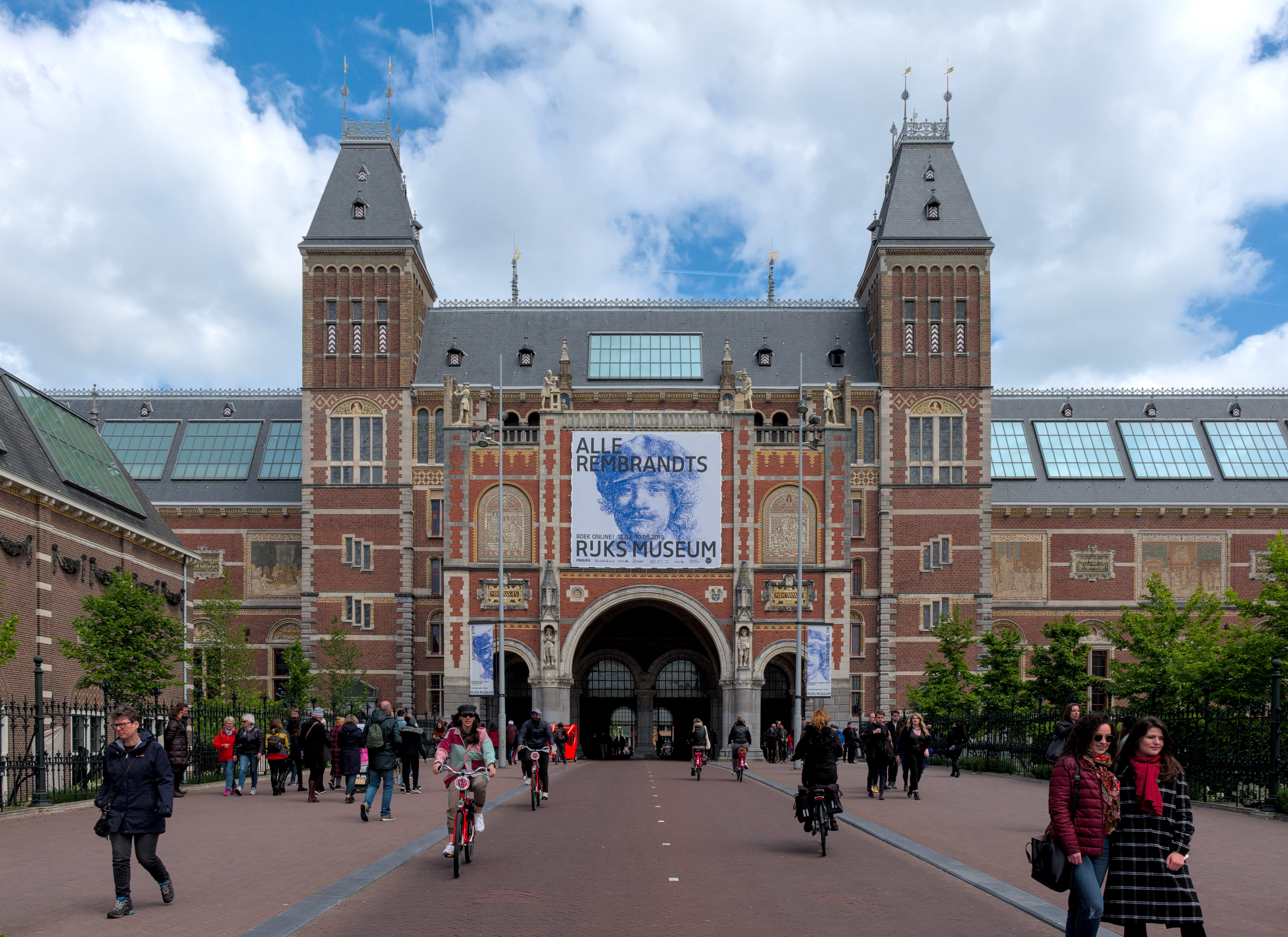 South facade of the Rijksmuseum with brick towers and a large entrance, people walking and cycling in front