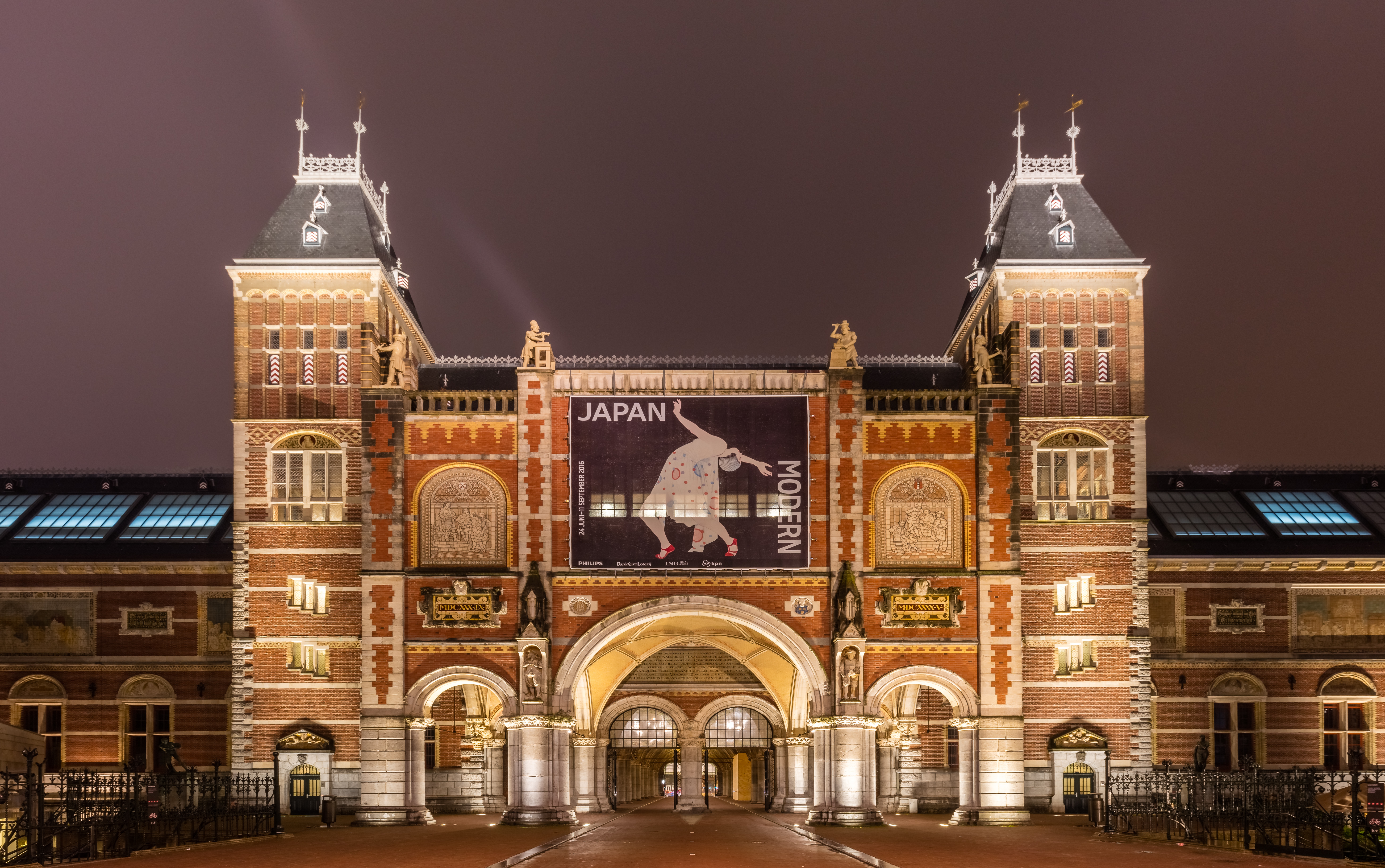 De gevel van het Rijksmuseum in de nacht, met een groot spandoek voor een Japan-tentoonstelling, verlichte torens en een boogvormige ingang.