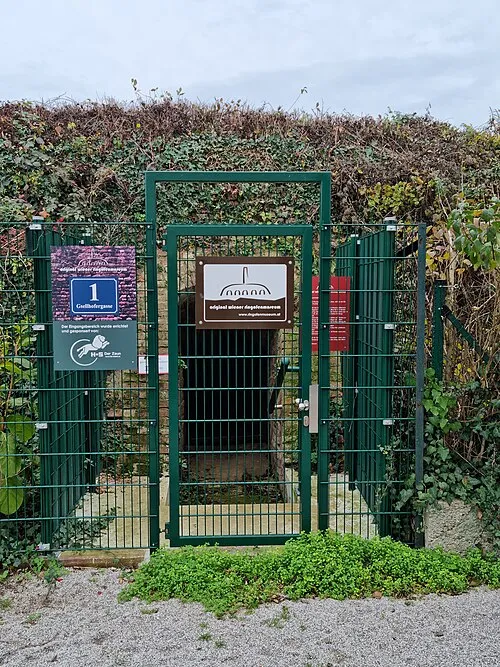 Zielona metalowa brama z tabliczkami Muzeum Ring Kiln, otoczona ceglanym murem i zarośniętą roślinnością.