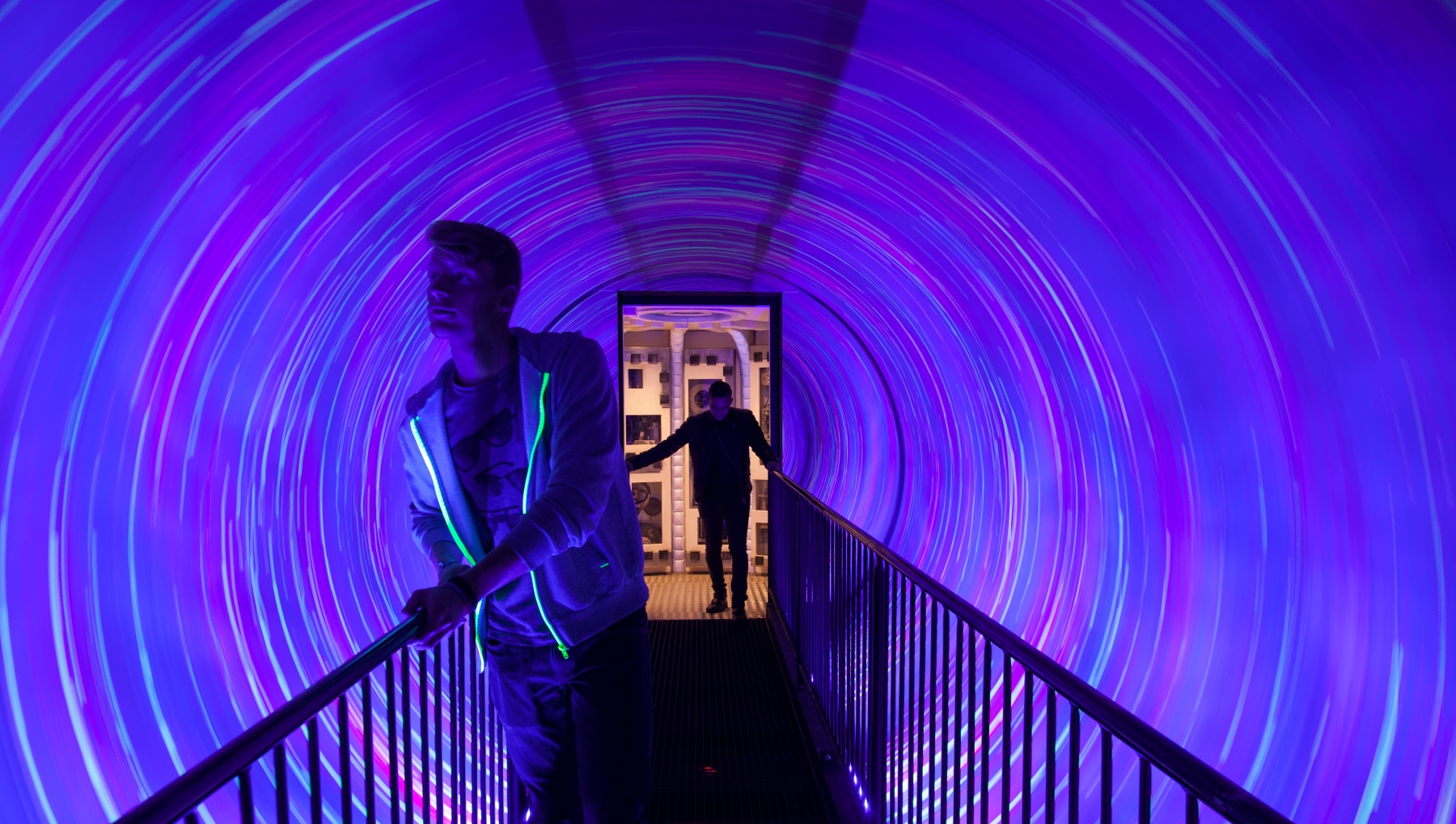 Dos personas caminan a través de una vibrante exposición de túnel de luz con patrones de neón púrpuras y azules arremolinados, se ve una escalera con barandillas negras